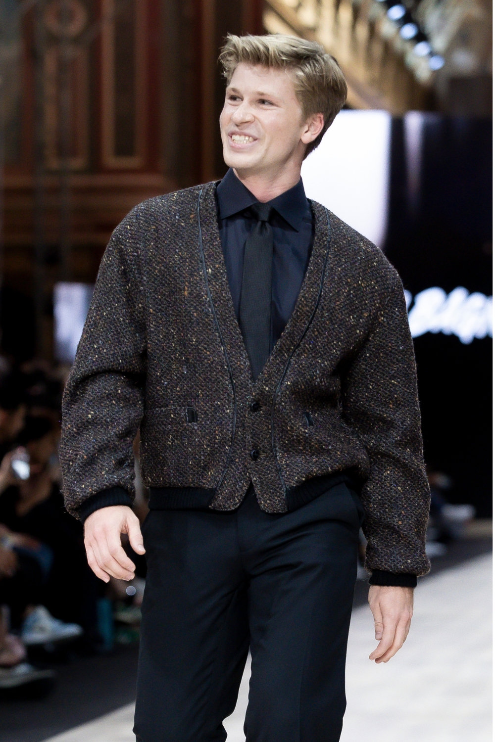 Robert Irwin wearing a grey cardigan, black shirt and tie walking a fashion runway.