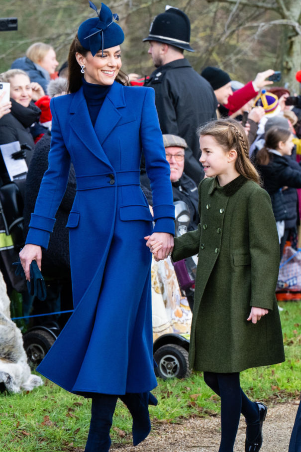 A woman in a blue coat and hat holds hands with a young girl in a green coat, smiling at an outdoor event.