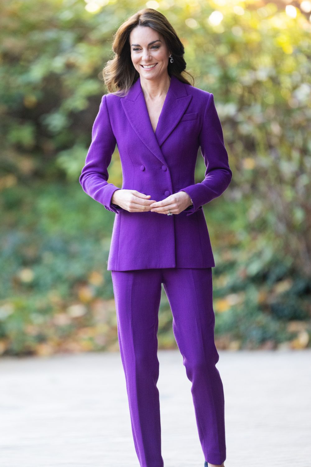 Woman in a vibrant purple suit stands outdoors, smiling, with a blurred leafy background.