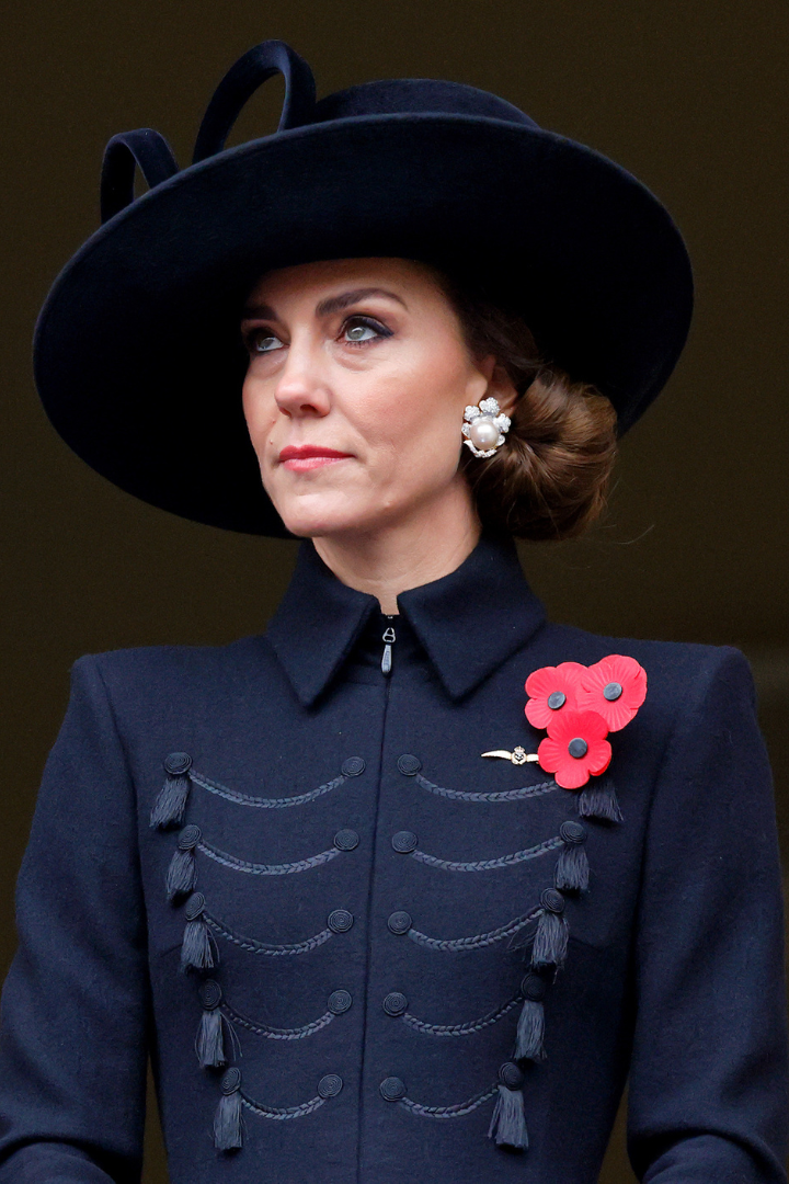 A woman in a dark coat and wide-brimmed hat with poppy brooches and pearl earrings.