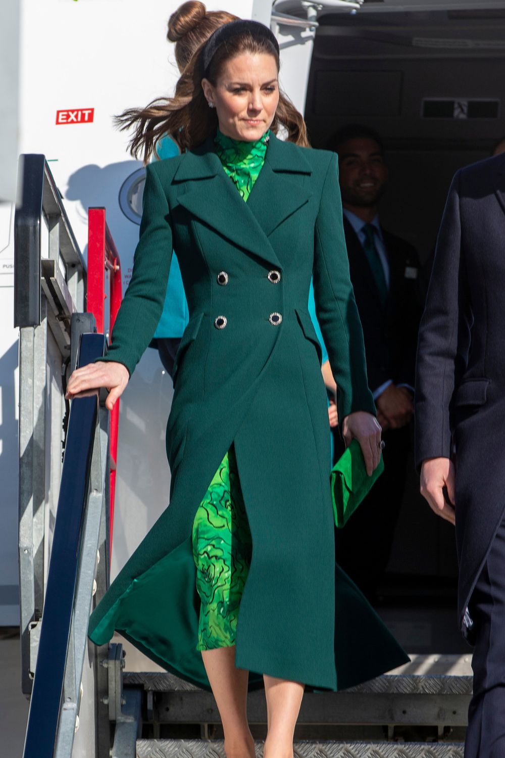 A woman in a dark green coat with a patterned dress exits an airplane, holding a matching green clutch.