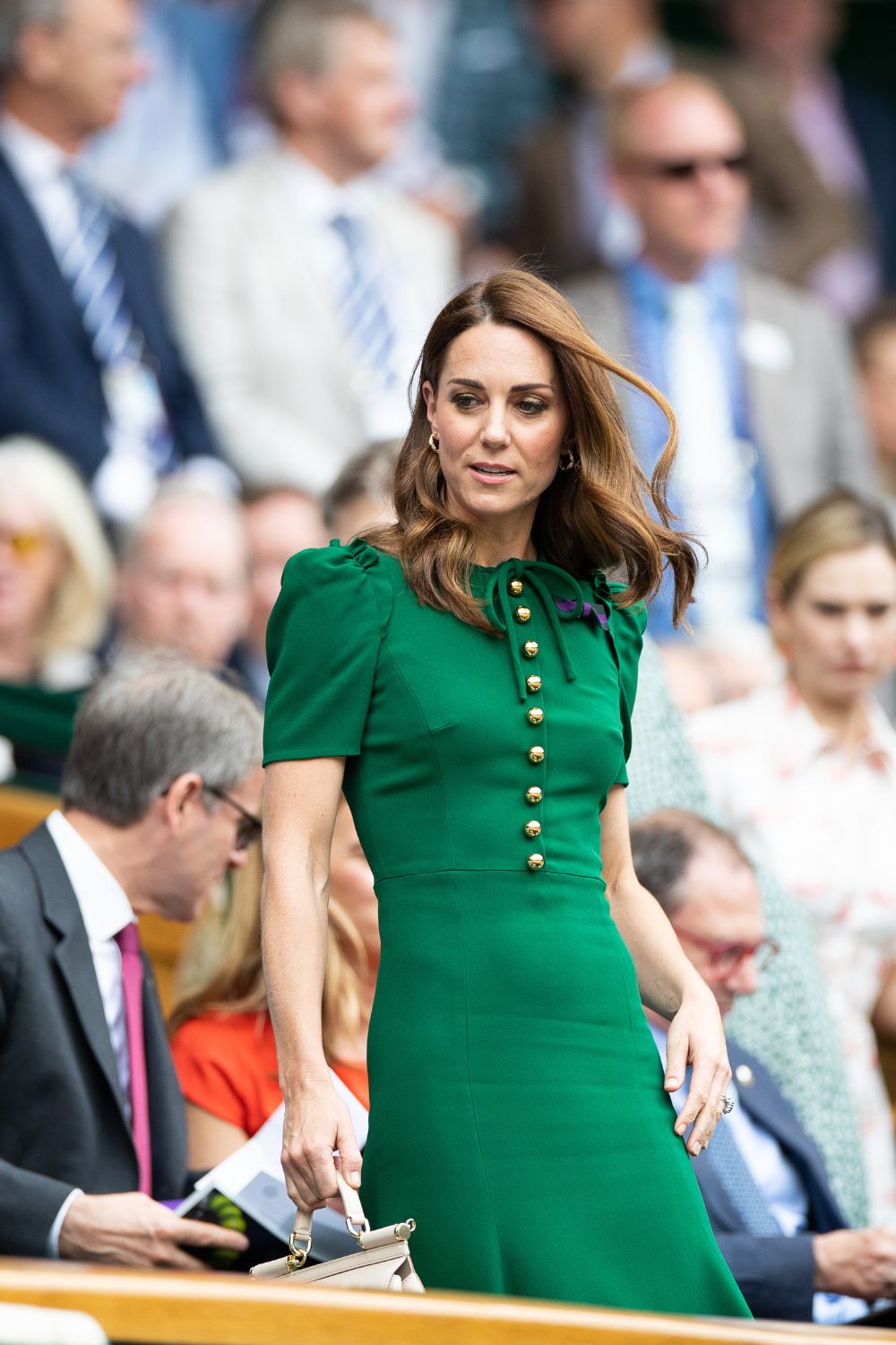 A woman in a green dress with gold buttons stands holding a cream handbag, surrounded by a seated crowd.