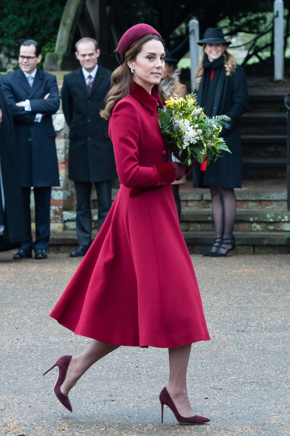 A woman in a red coat and hat, holding a bouquet, walks outdoors with people in the background.