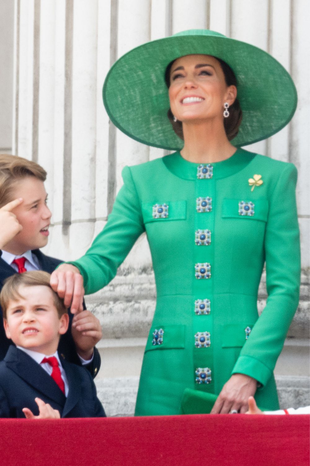 A woman in a green dress and hat smiles beside two children in suits, standing at a railing in front of a building.
