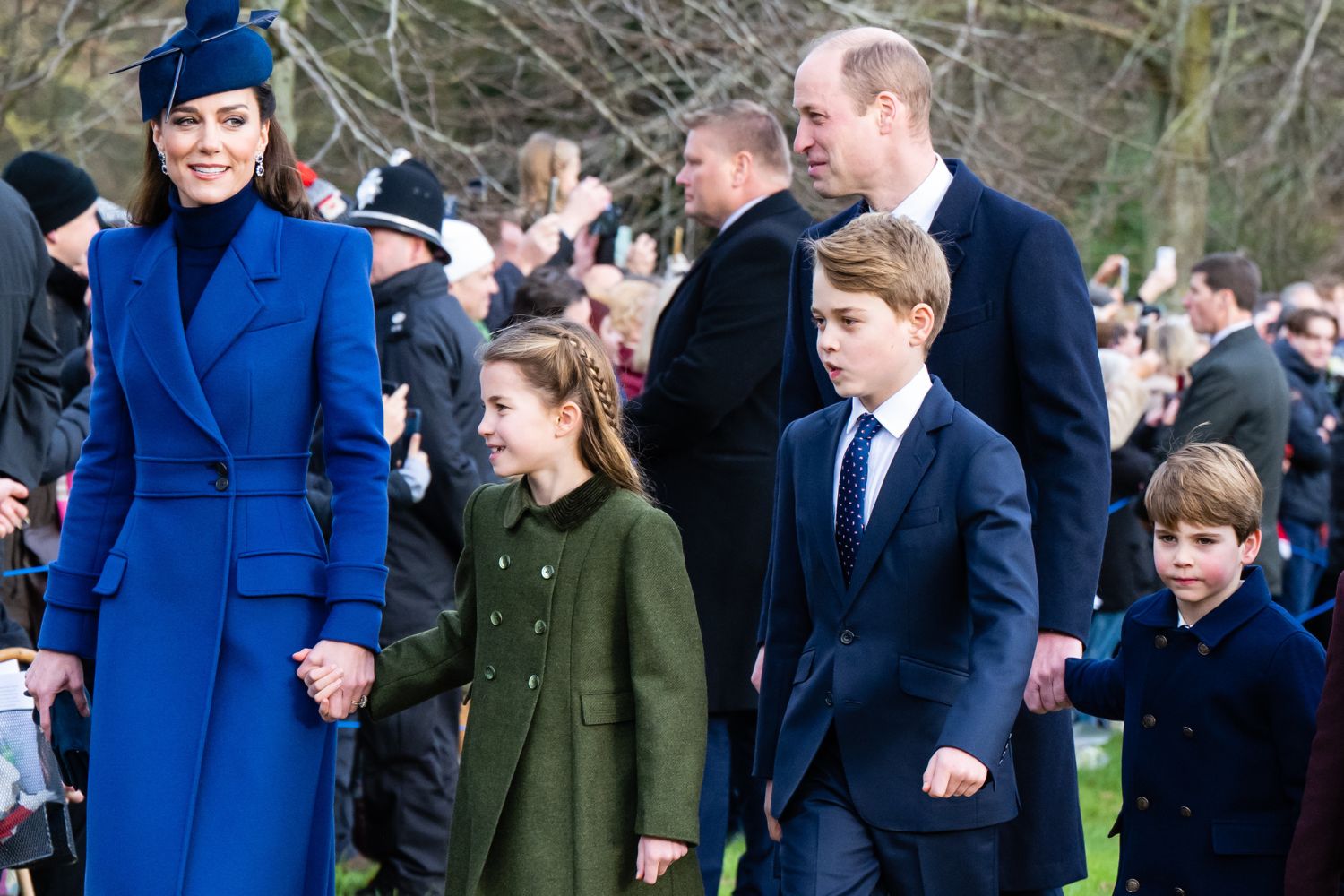 A family walking outdoors, dressed in formal winter attire, with a crowd in the background.