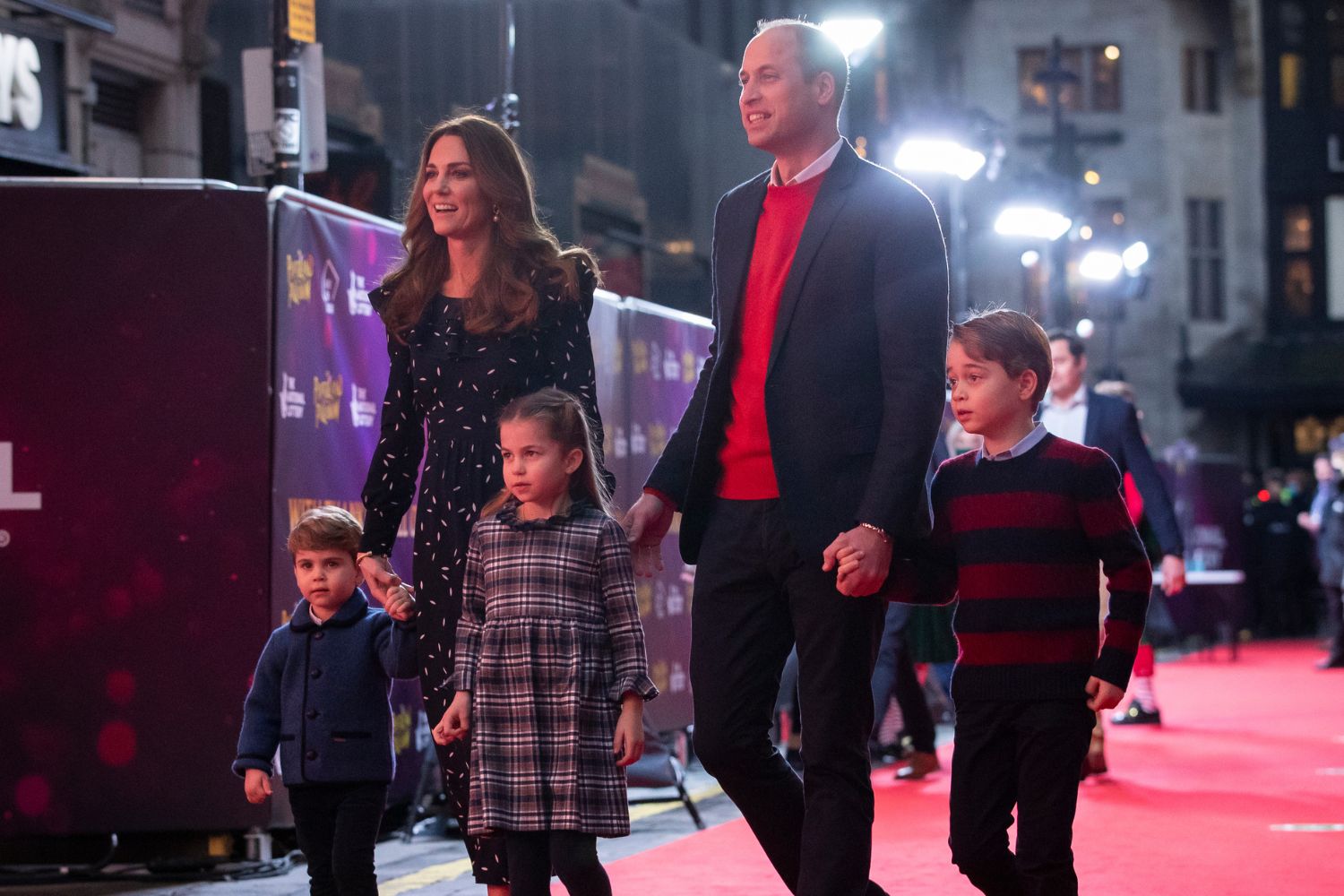 A family walking on a red carpet, holding hands, with bright lights in the background.