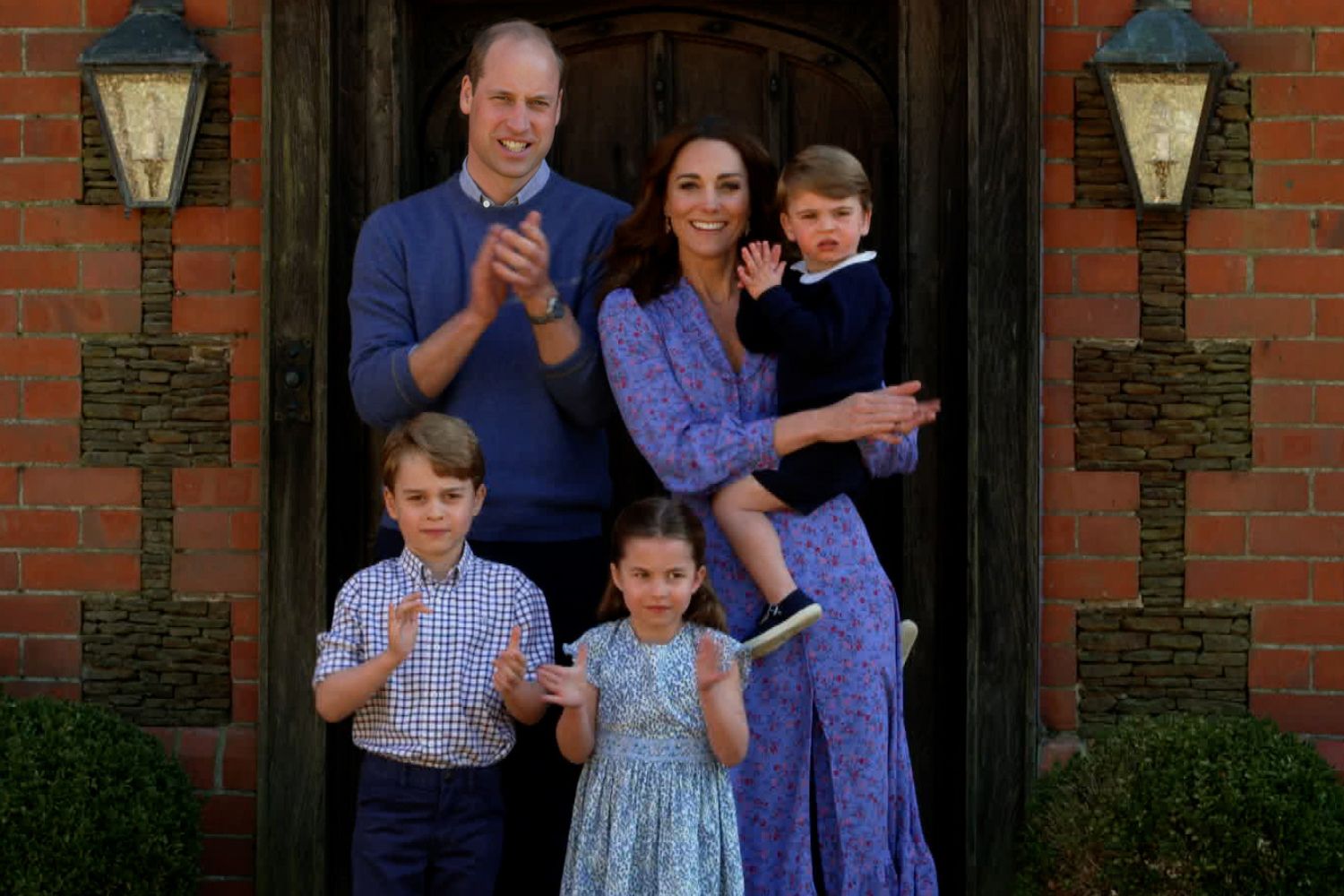A family of five clapping outside a brick house, with a man, woman, and three children.
