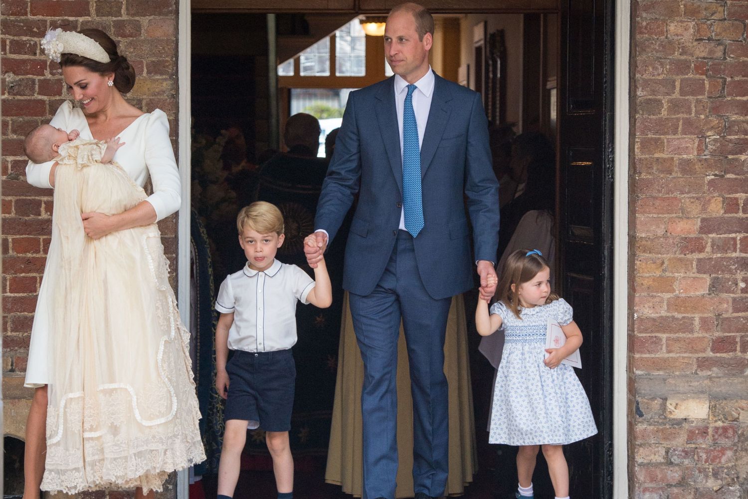 Family exiting church; woman holding baby, man in suit holding boy and girl's hands, brick backdrop.