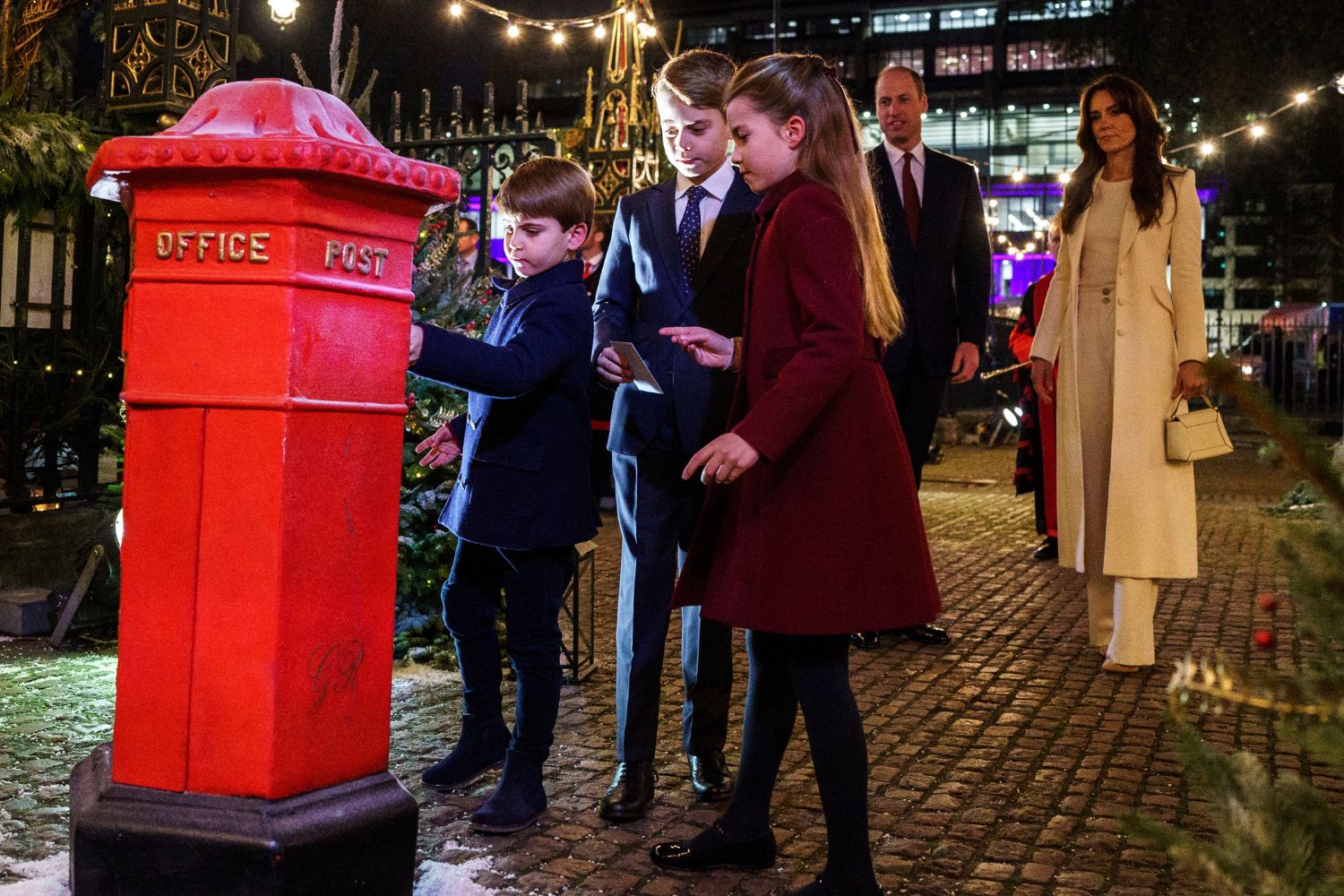 Children post letters in a red postbox outdoors, with adults watching, during a festive evening event.