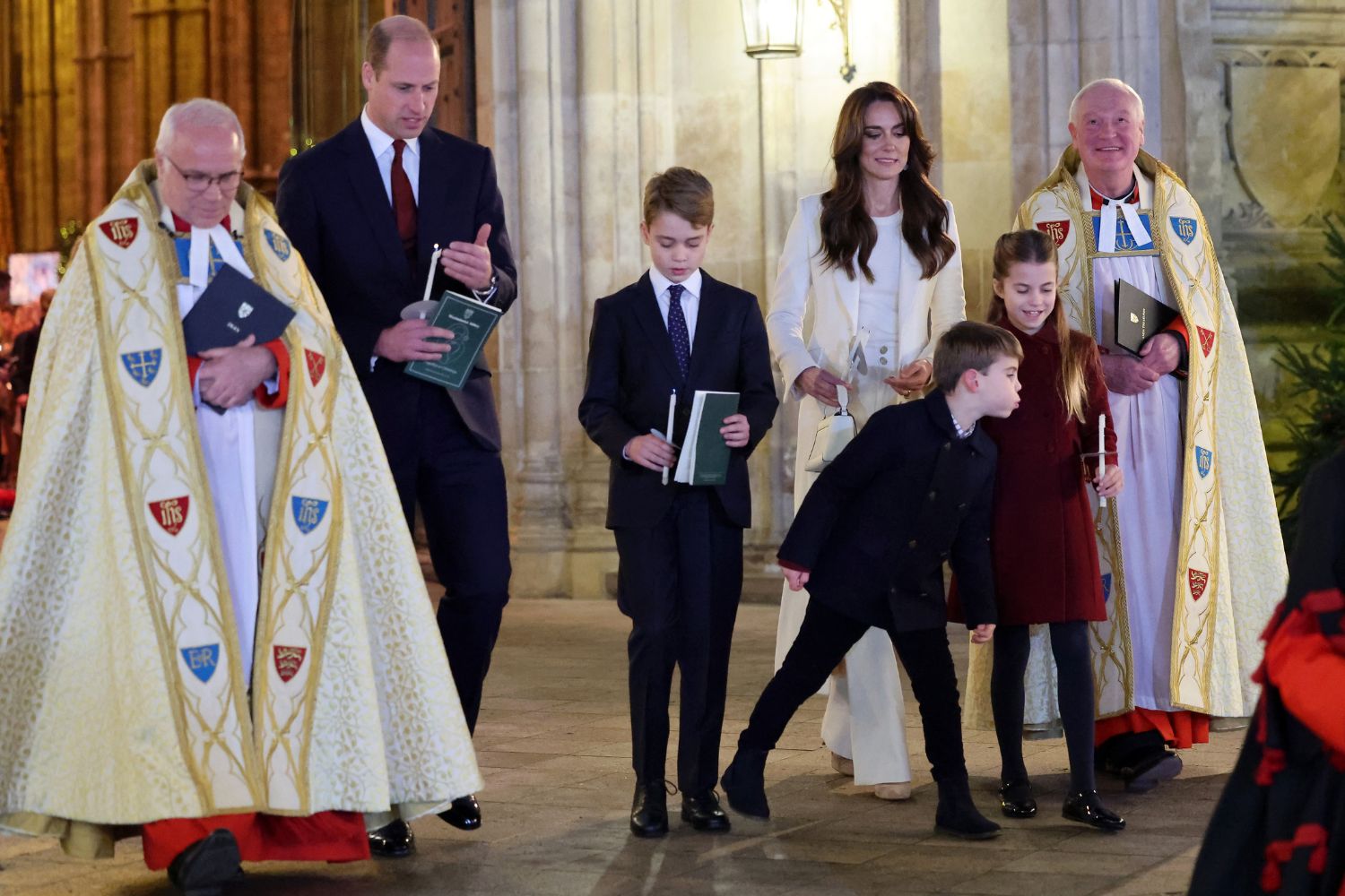 Royal family with clergy holding candles and programs in a church setting, dressed formally, two children interacting.