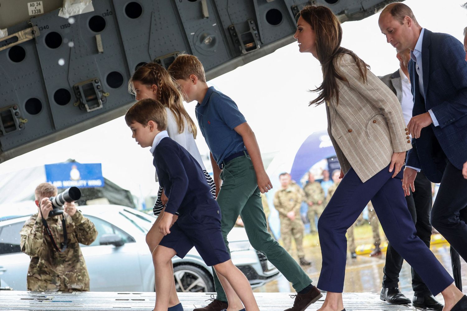 Family boarding an aircraft as a photographer captures the moment; military personnel are visible in the background.