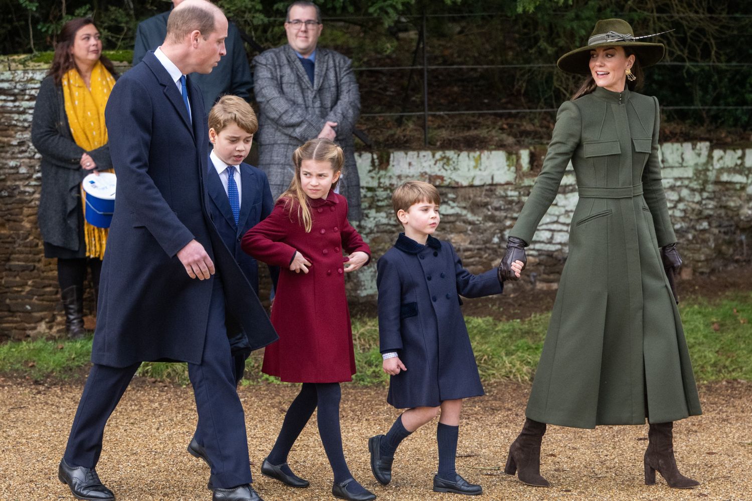 Royal family walking, with woman in green coat holding hands with young boy in blue coat, and man leading other children.