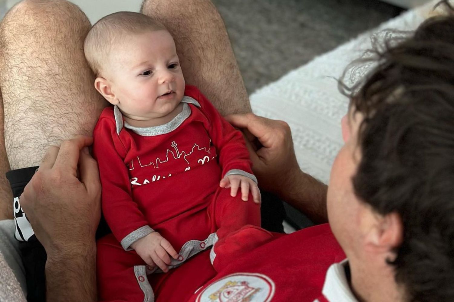Baby dressed in red sitting on a person's lap, wearing a top with city skyline print.