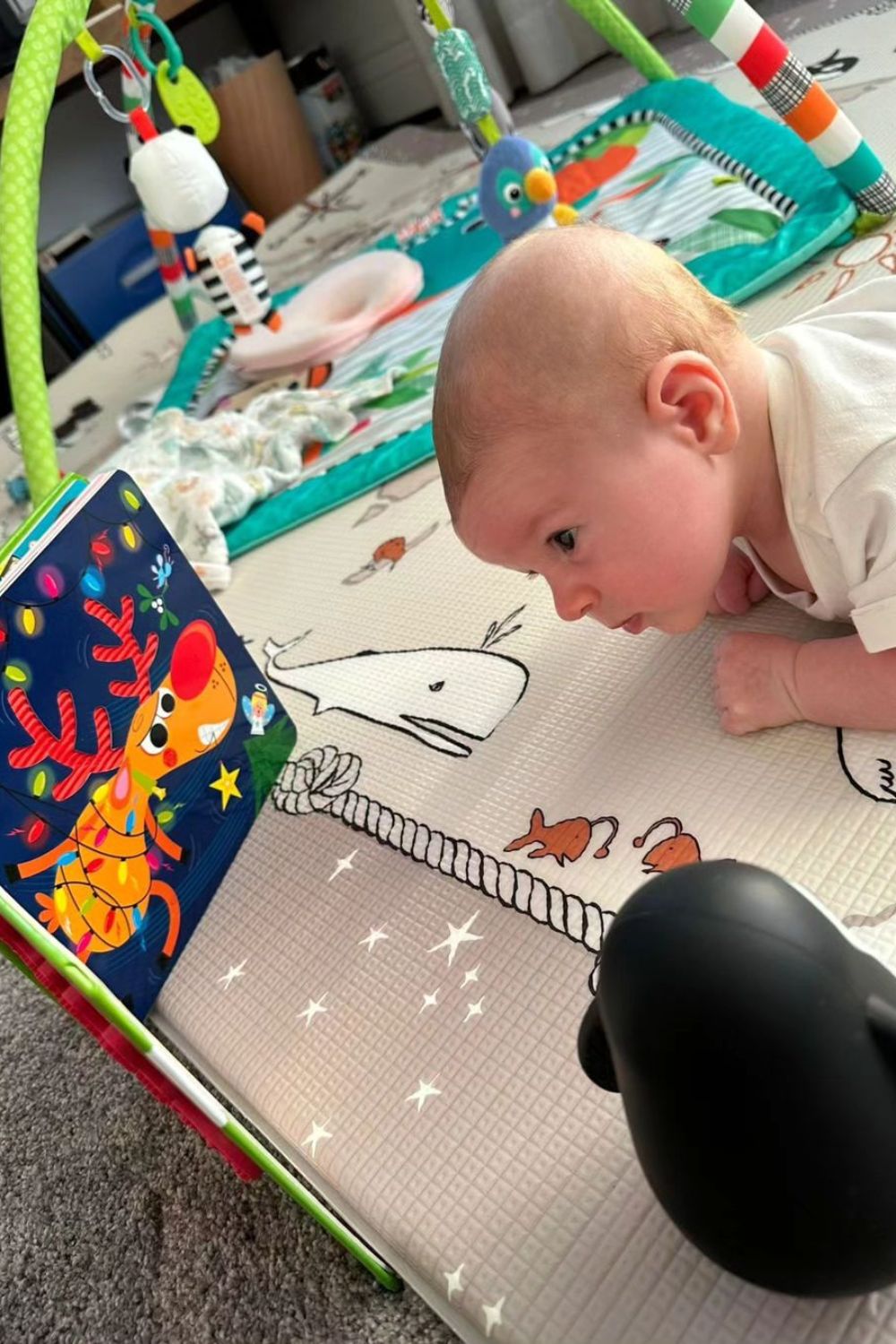 Baby lying on a play mat with a reindeer book and toys nearby.