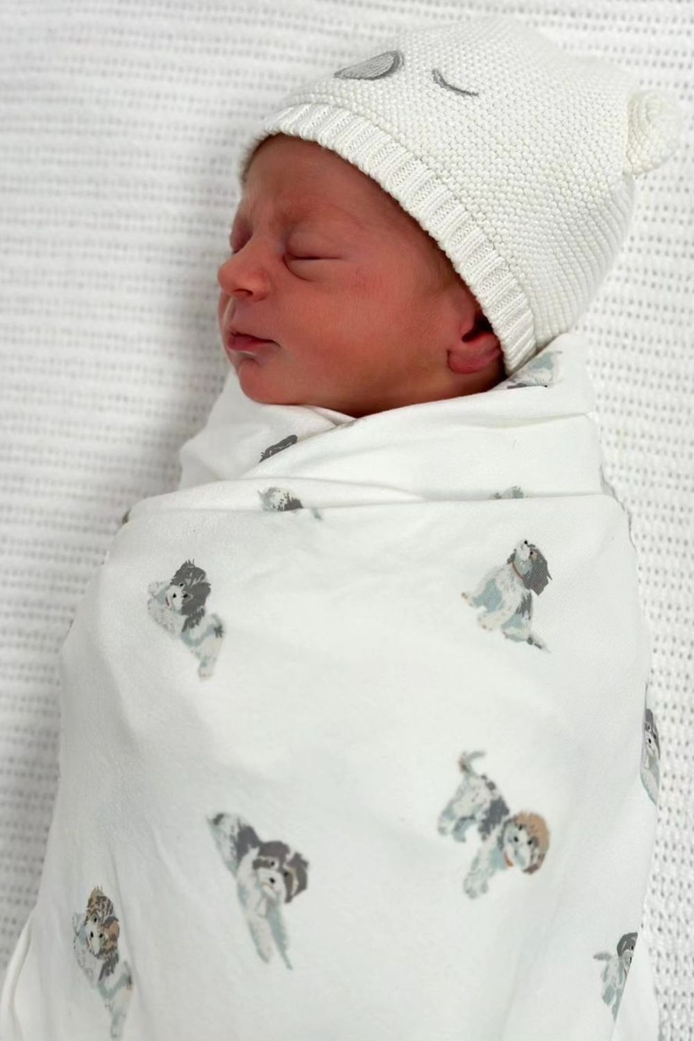 Newborn baby wrapped in a white blanket with teddy prints, wearing a knitted hat with bear ears, sleeping peacefully.