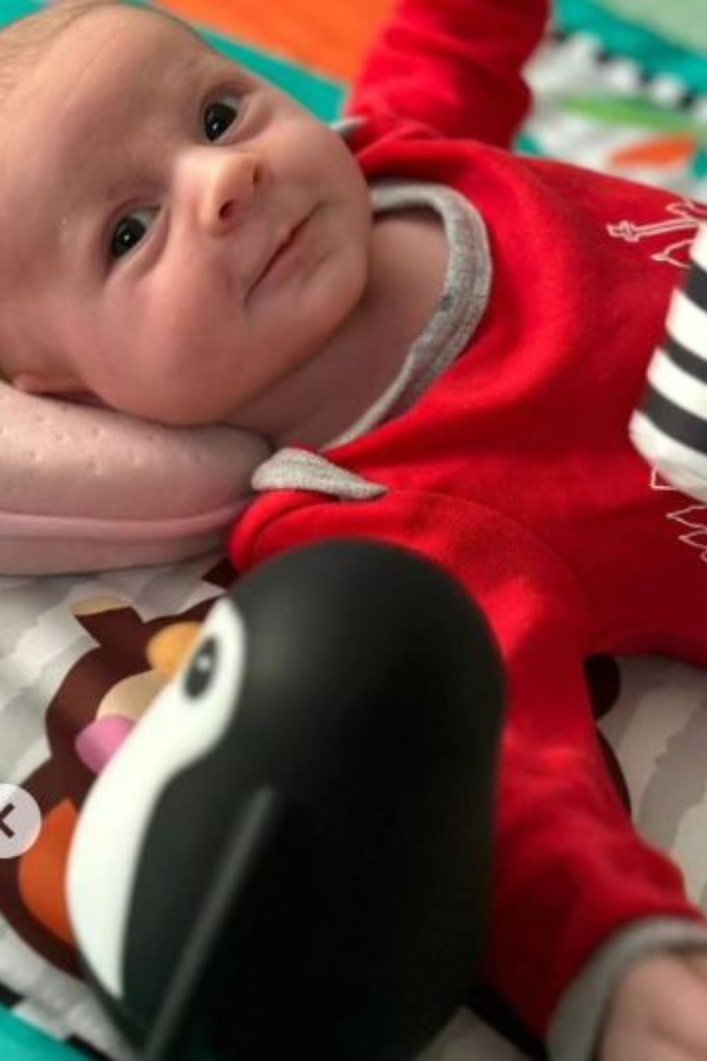 Baby wearing a red outfit, lying on a play mat, looks at a penguin toy in the foreground.