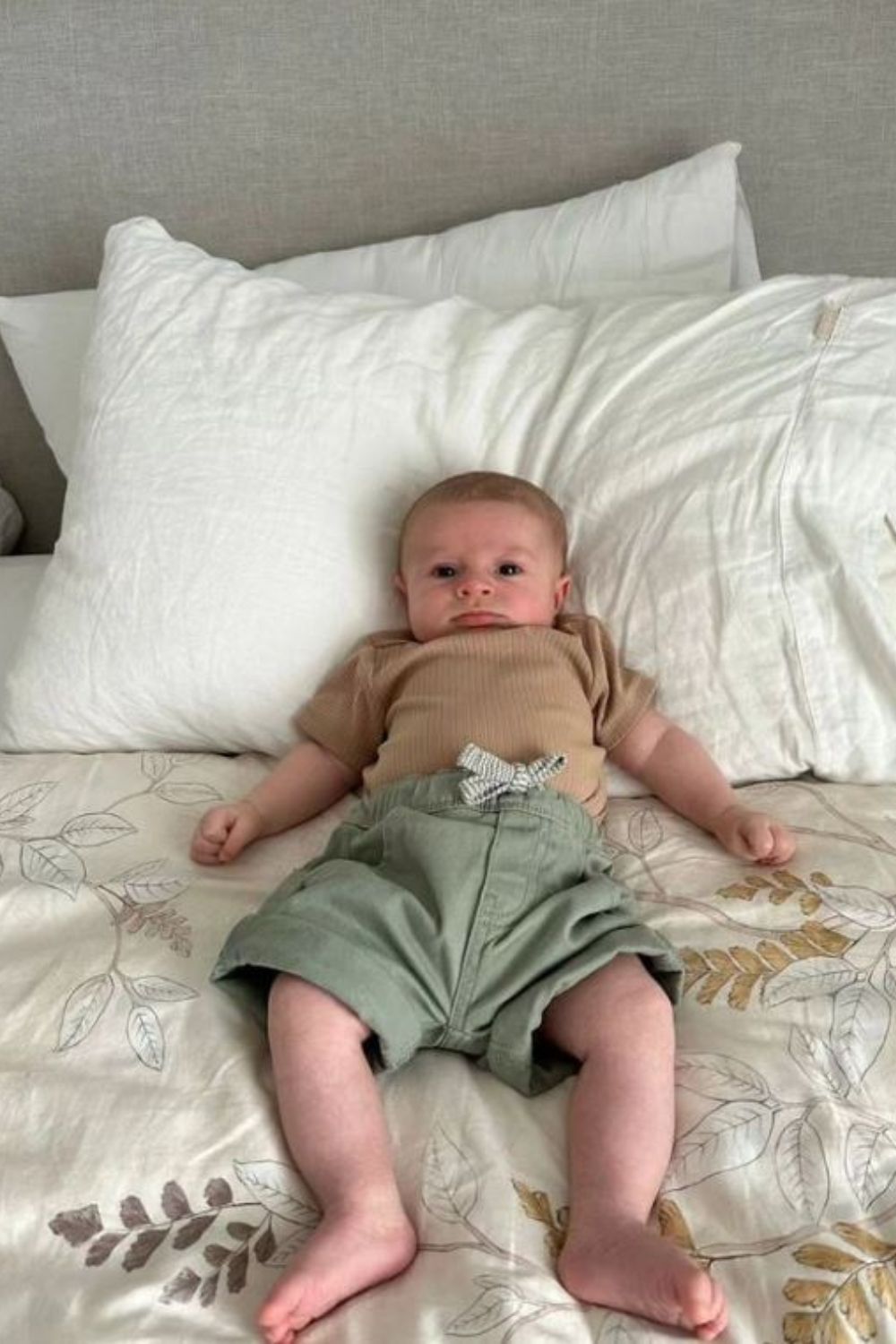 Baby lying on a bed with white pillows, wearing a beige shirt and green shorts, looking at the camera.