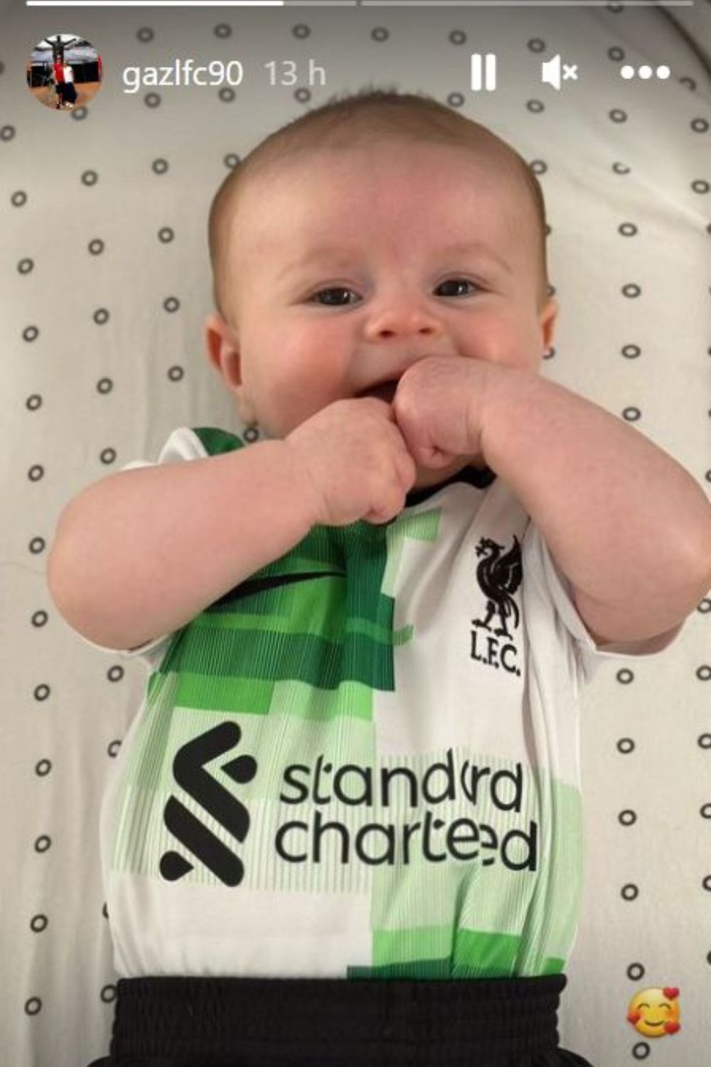 Baby wearing a Liverpool FC onesie, lying on a patterned blanket with hands near mouth.