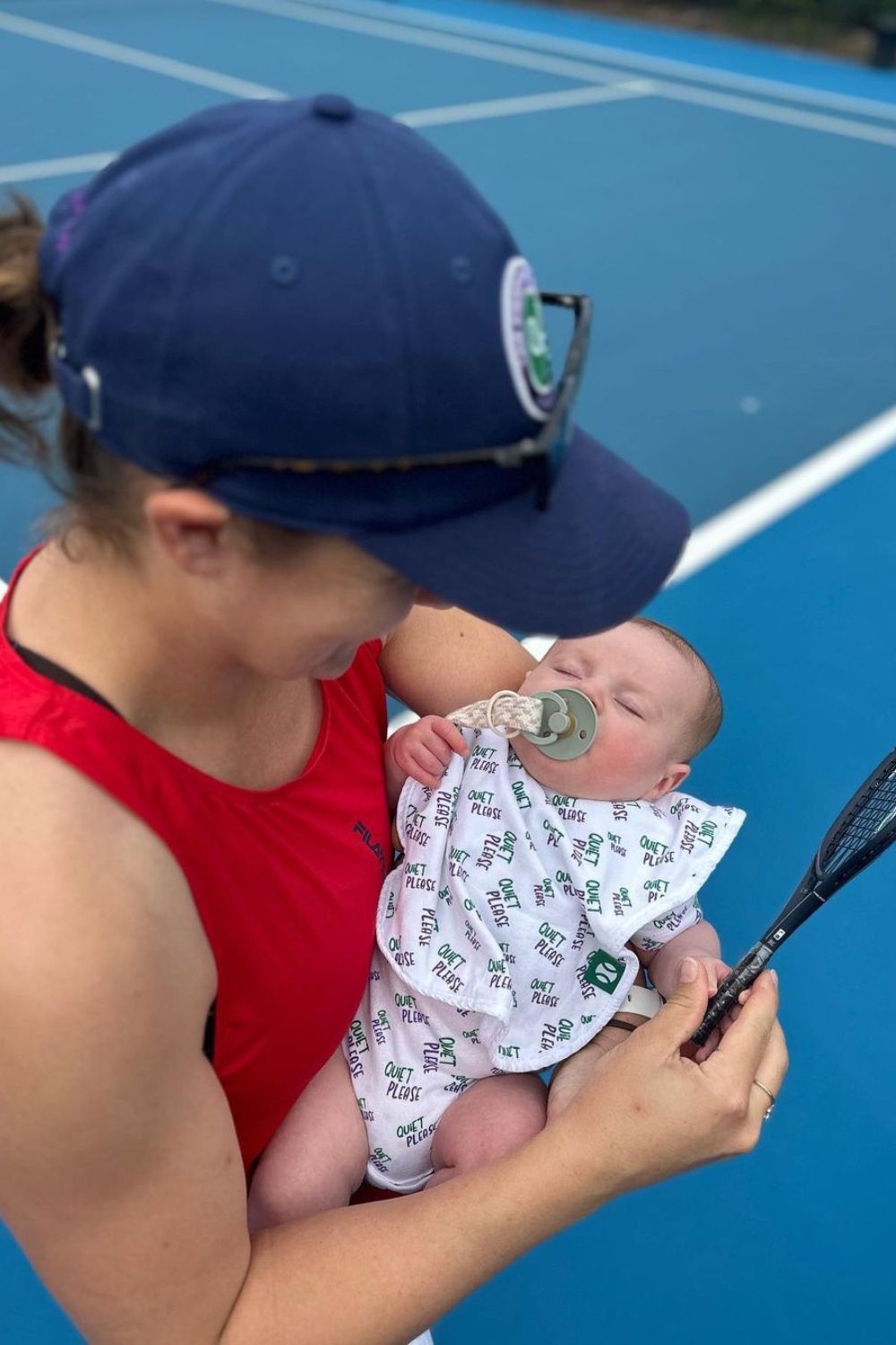 A woman in a red top, wearing a blue cap, holds a baby with a pacifier and "Quiet Please" bib on a tennis court.