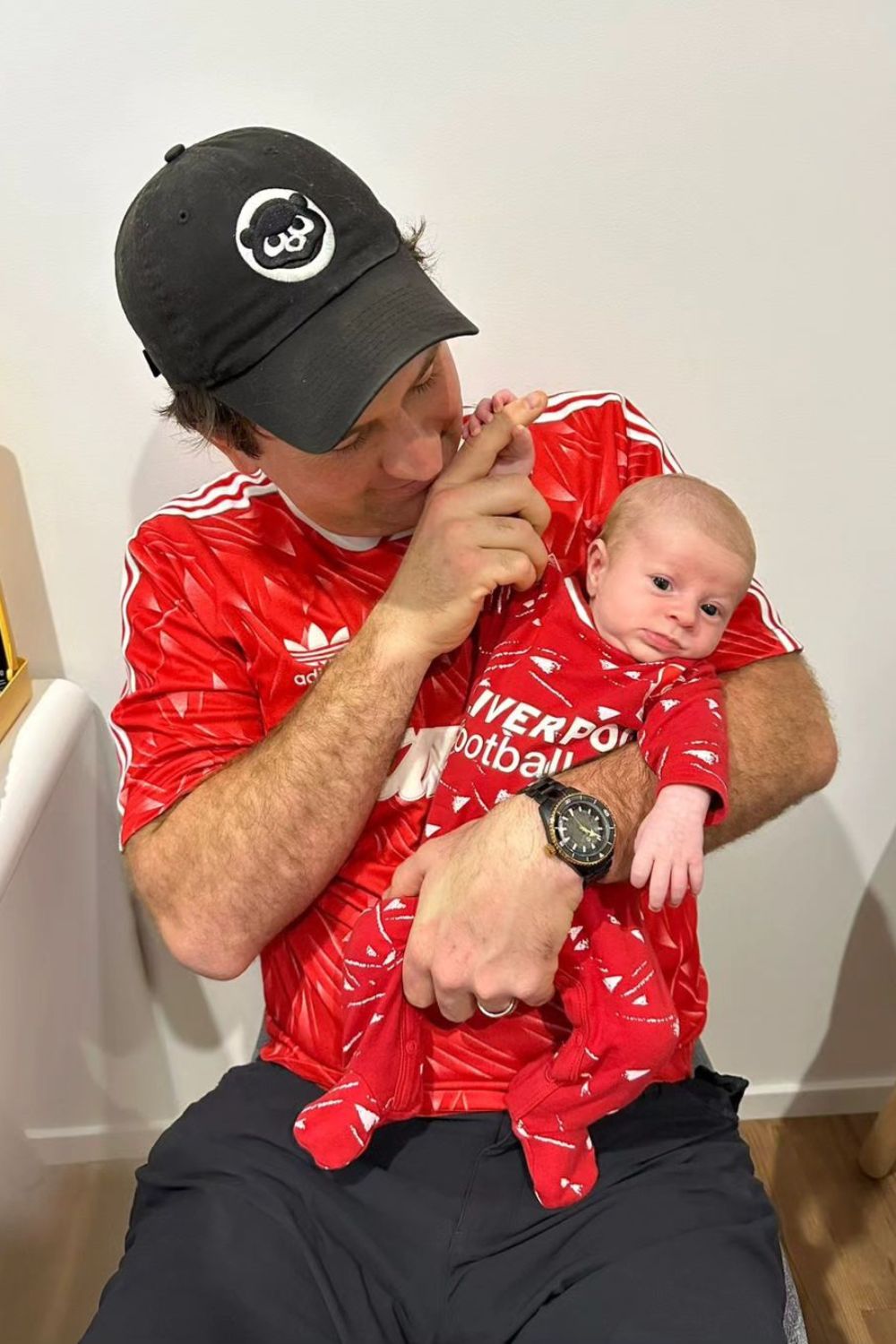 Man in red sports jersey holding a baby dressed in matching outfit, wearing a black cap and watch.