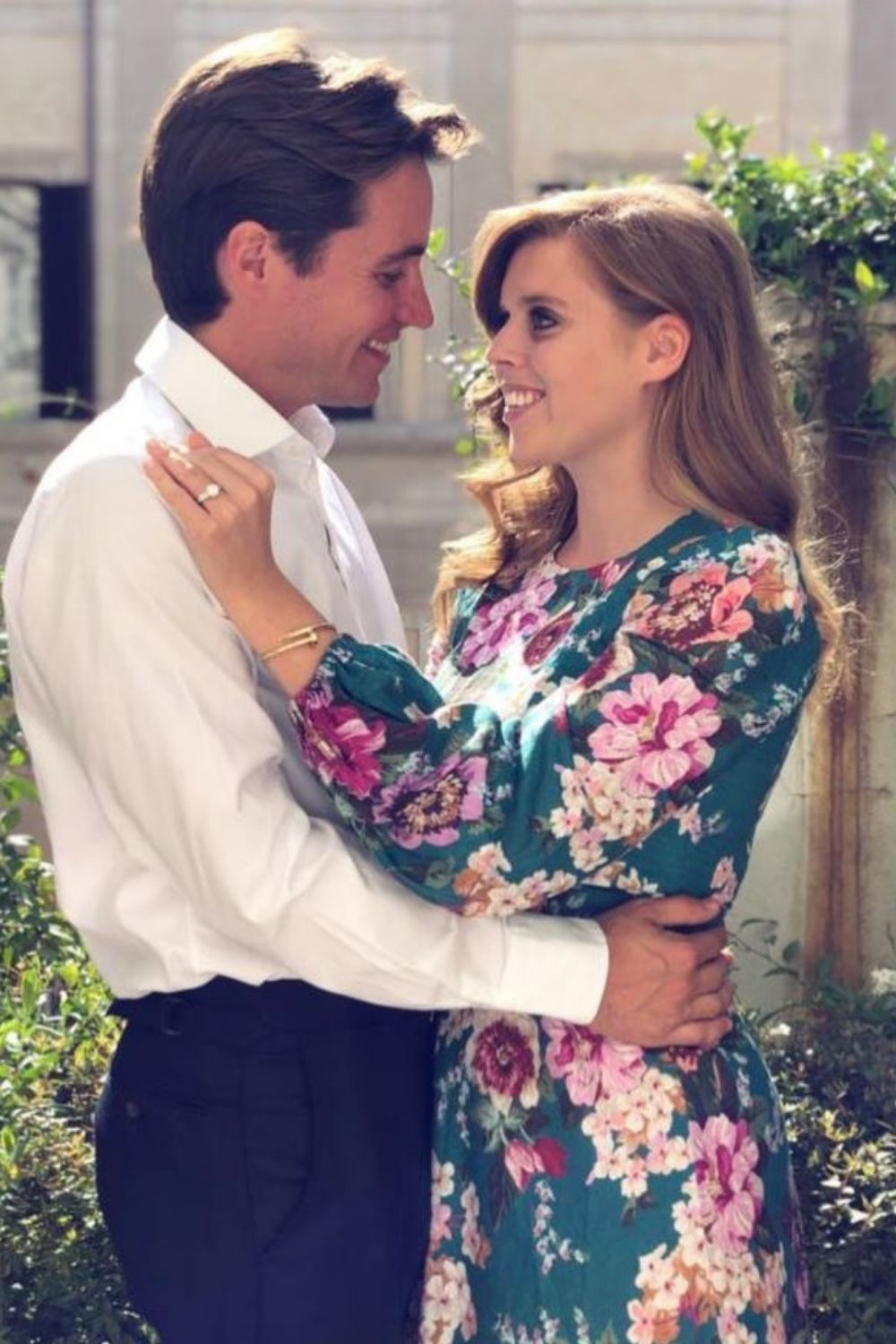 A couple embraces outdoors; the woman wears a floral dress, the man in a white shirt smiles at her.