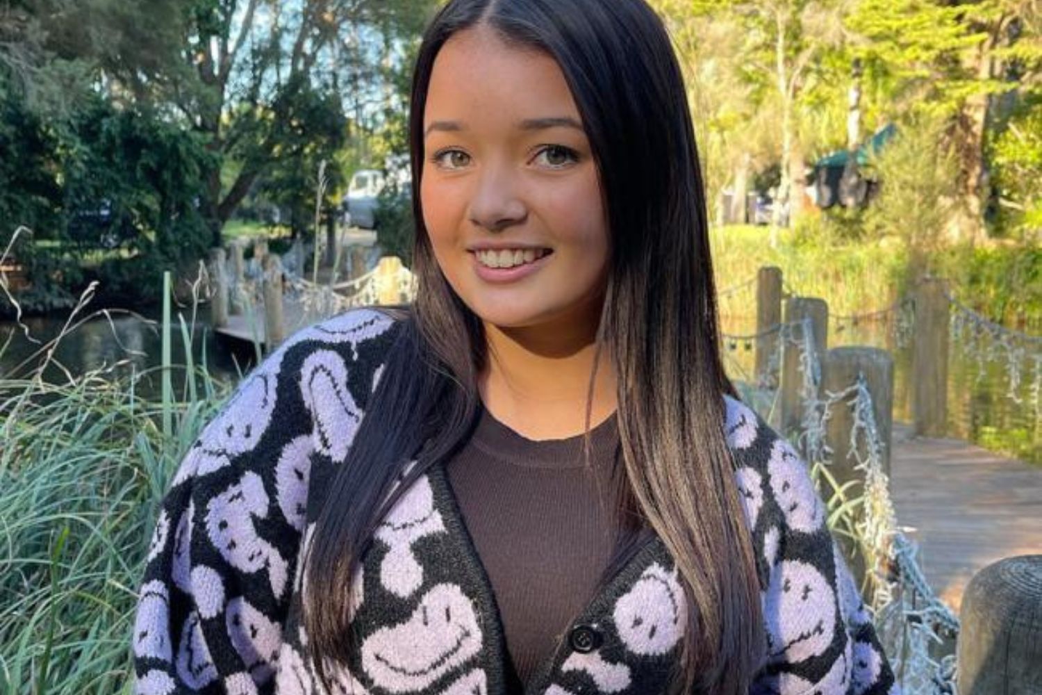 A person with long dark hair smiling, wearing a sweater with smiley faces, standing on a wooden bridge in a lush park.