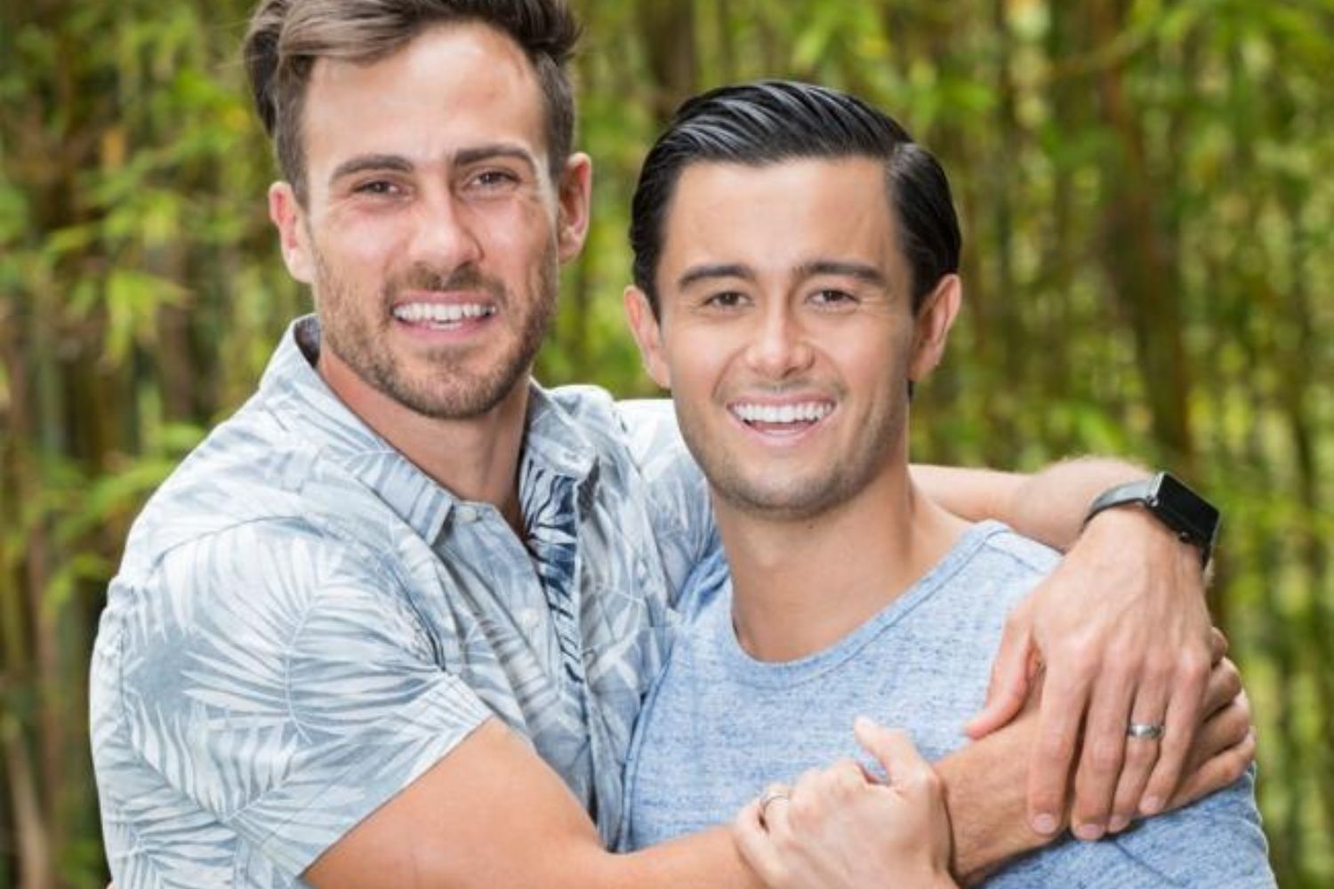 Two smiling men embrace outdoors, with greenery in the background, one wearing a tropical shirt and the other a gray t-shirt.