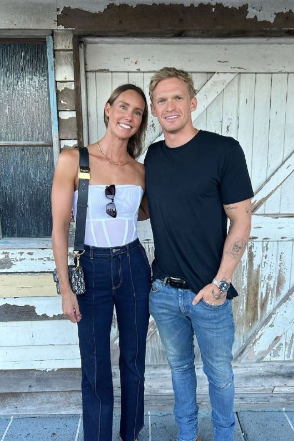 Two people smiling and posing together in casual outfits, standing in front of a rustic wooden door.