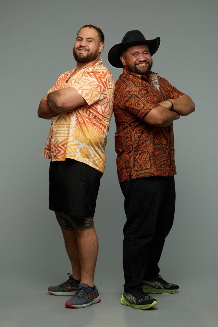 Two men standing back to back, both smiling. One wears an orange shirt and shorts, the other an orange-patterned shirt and hat.