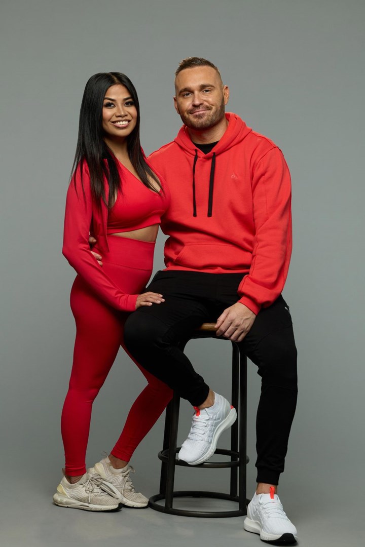 A woman in red and a man in a red hoodie sit closely together; both are smiling at the camera.