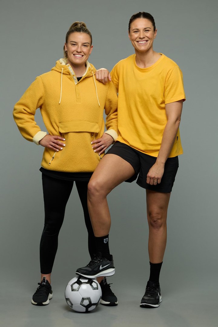 Two women in yellow tops smiling; one has a soccer ball under her foot.