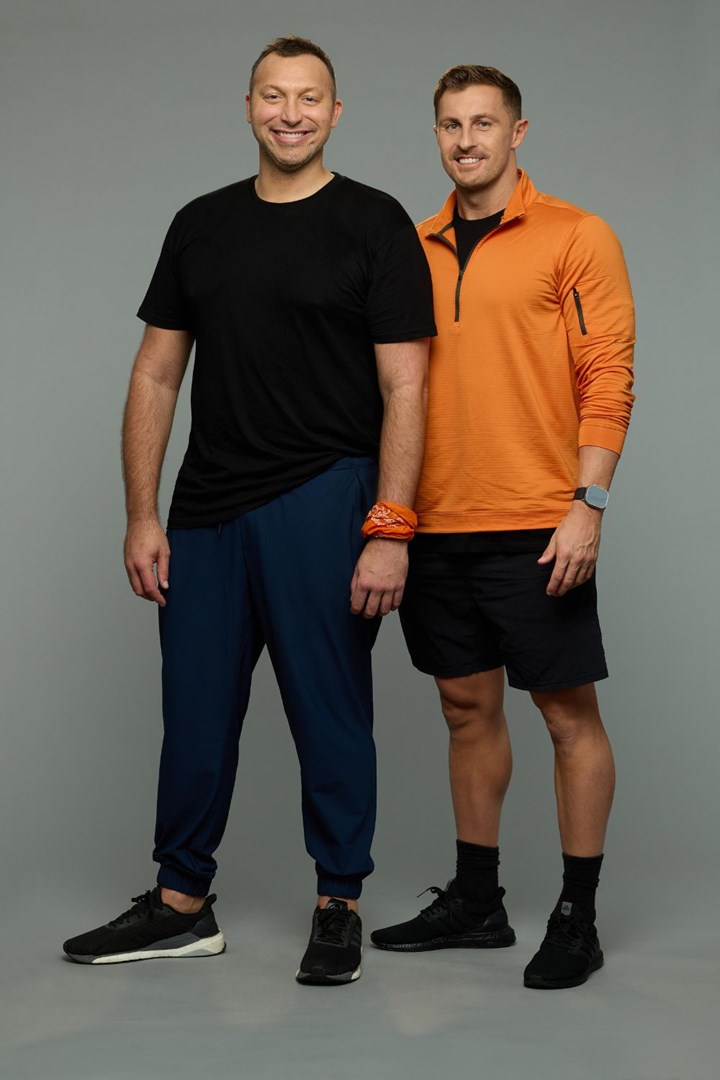 Two men standing side by side, smiling, one in a black outfit and the other in orange, against a plain background.