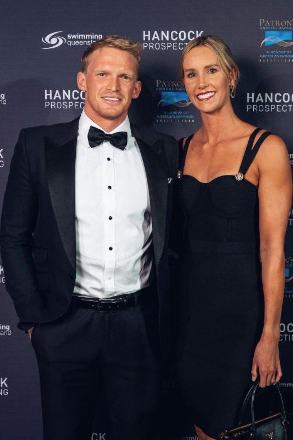 A man in a tuxedo and a woman in a black dress pose together on a red carpet event.