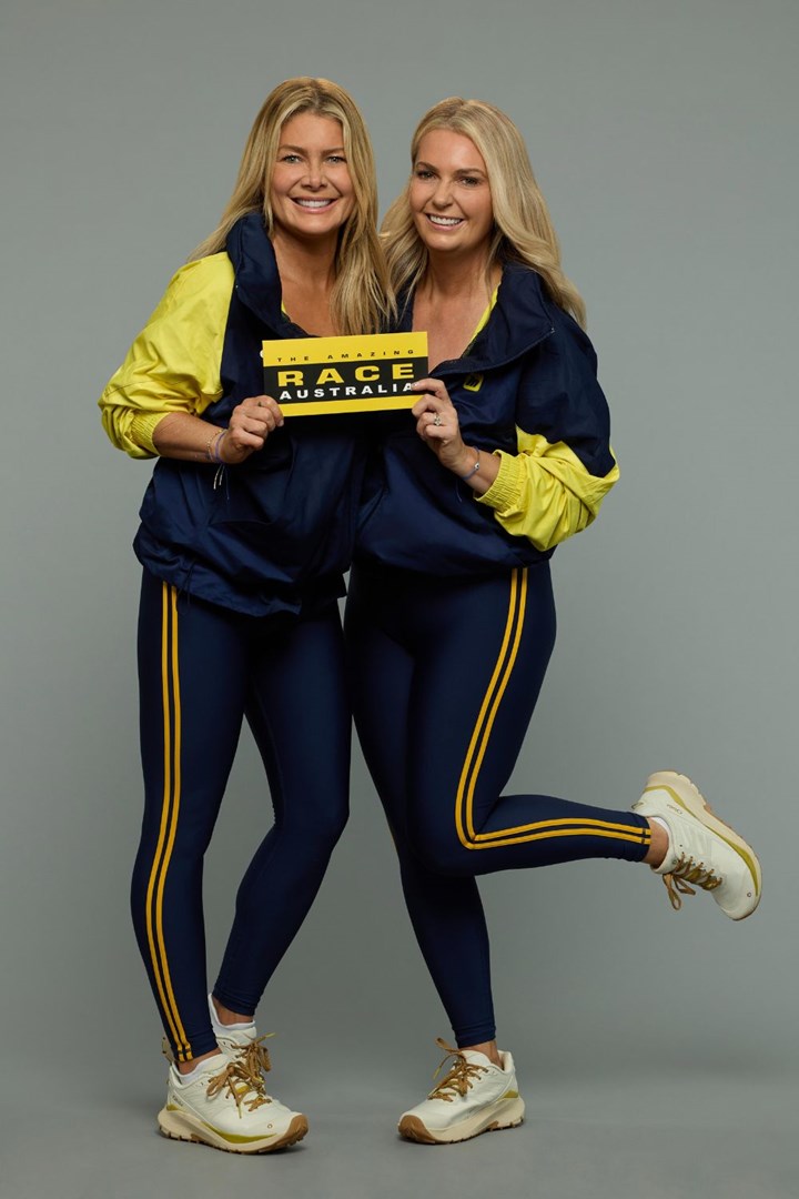 Two women in matching athletic outfits hold a "The Amazing Race Australia" sign, smiling against a gray background.