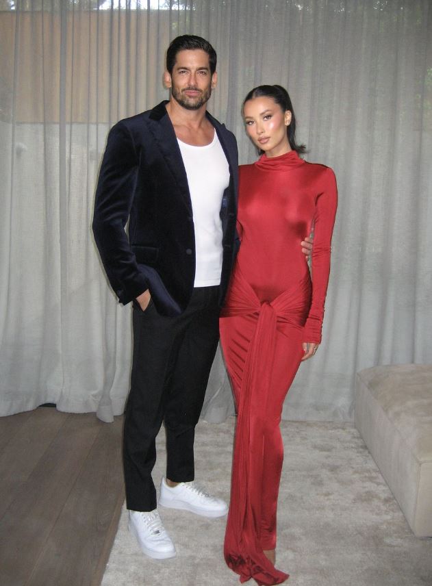 A man in a dark suit with a white shirt and a woman in a red dress stand together in a room with curtains.