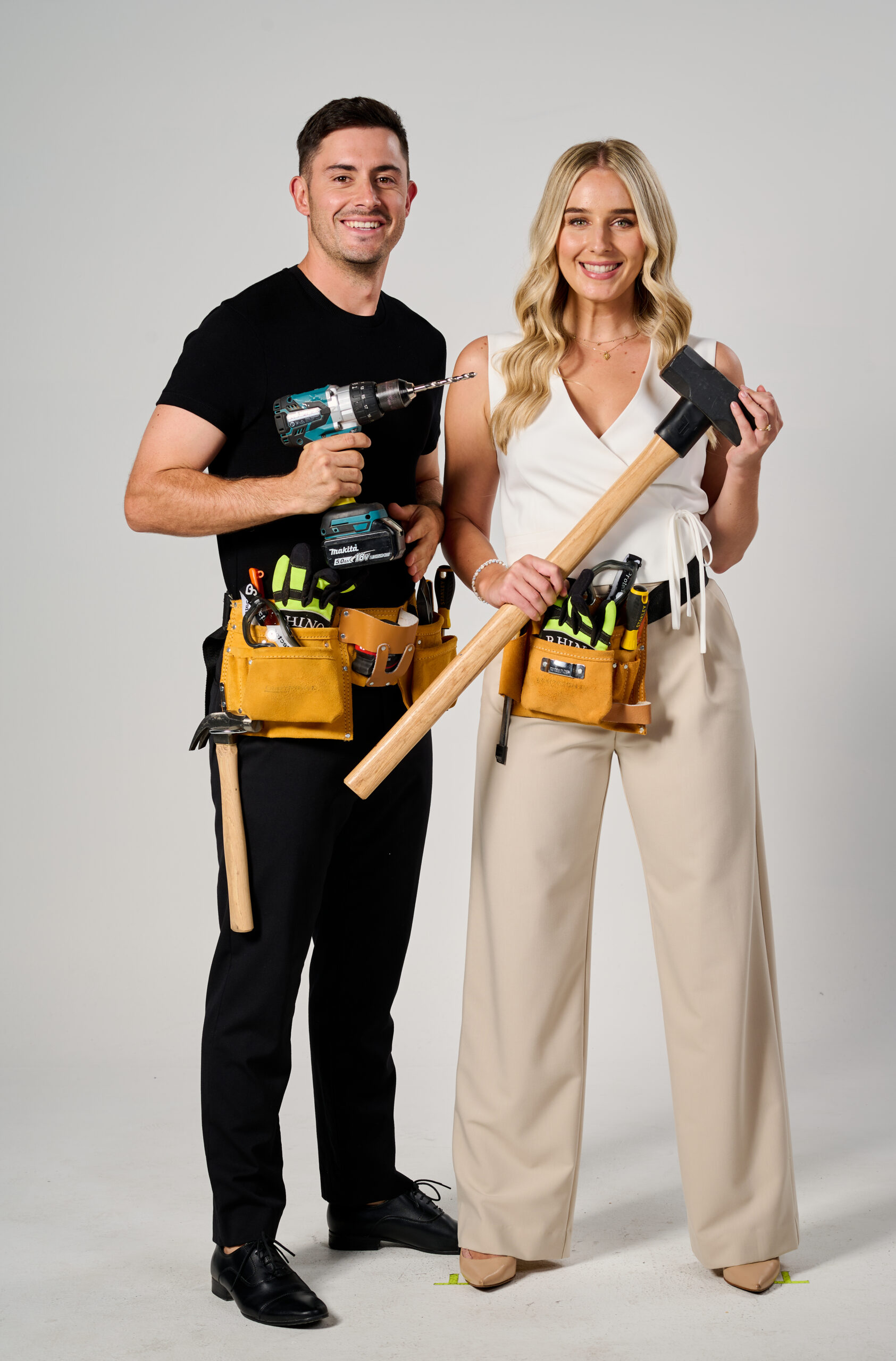 Couple posing with tools, man holding a drill and woman holding a mallet, both wearing tool belts.