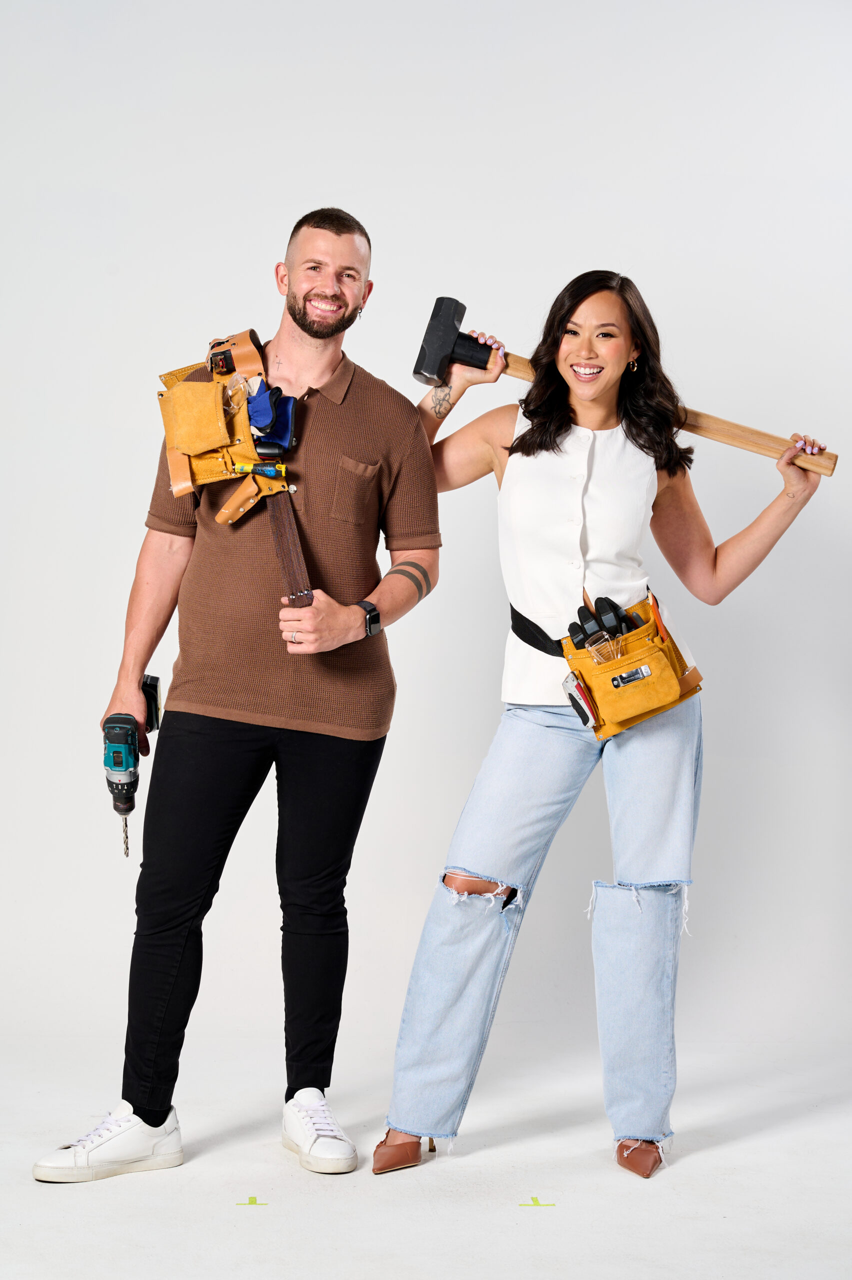 Man with drill and tool belt, standing next to woman holding a sledgehammer, both smiling in casual outfits.