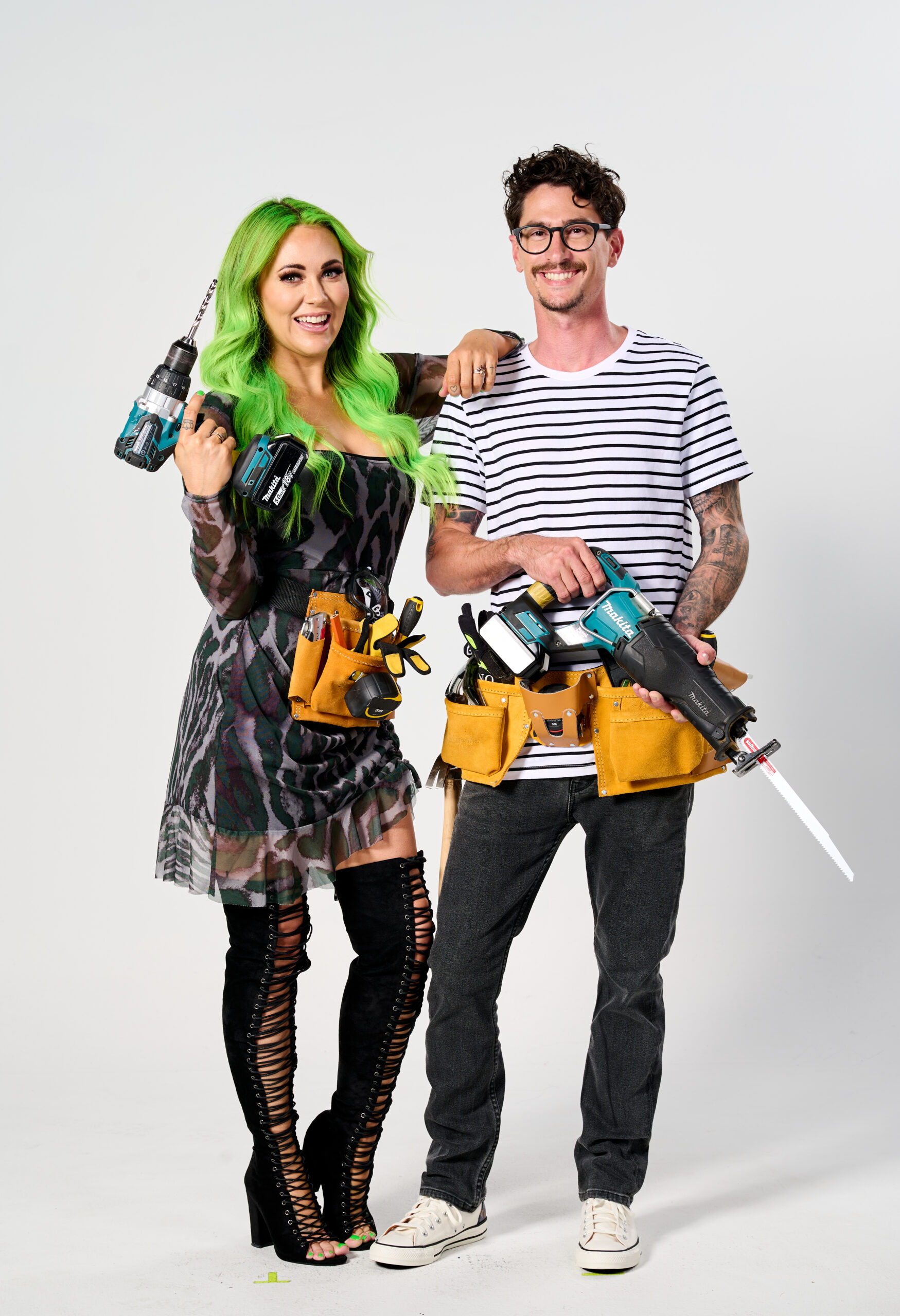 Two individuals pose with tools, one with bright green hair, wearing tool belts, conveying readiness for a building task.