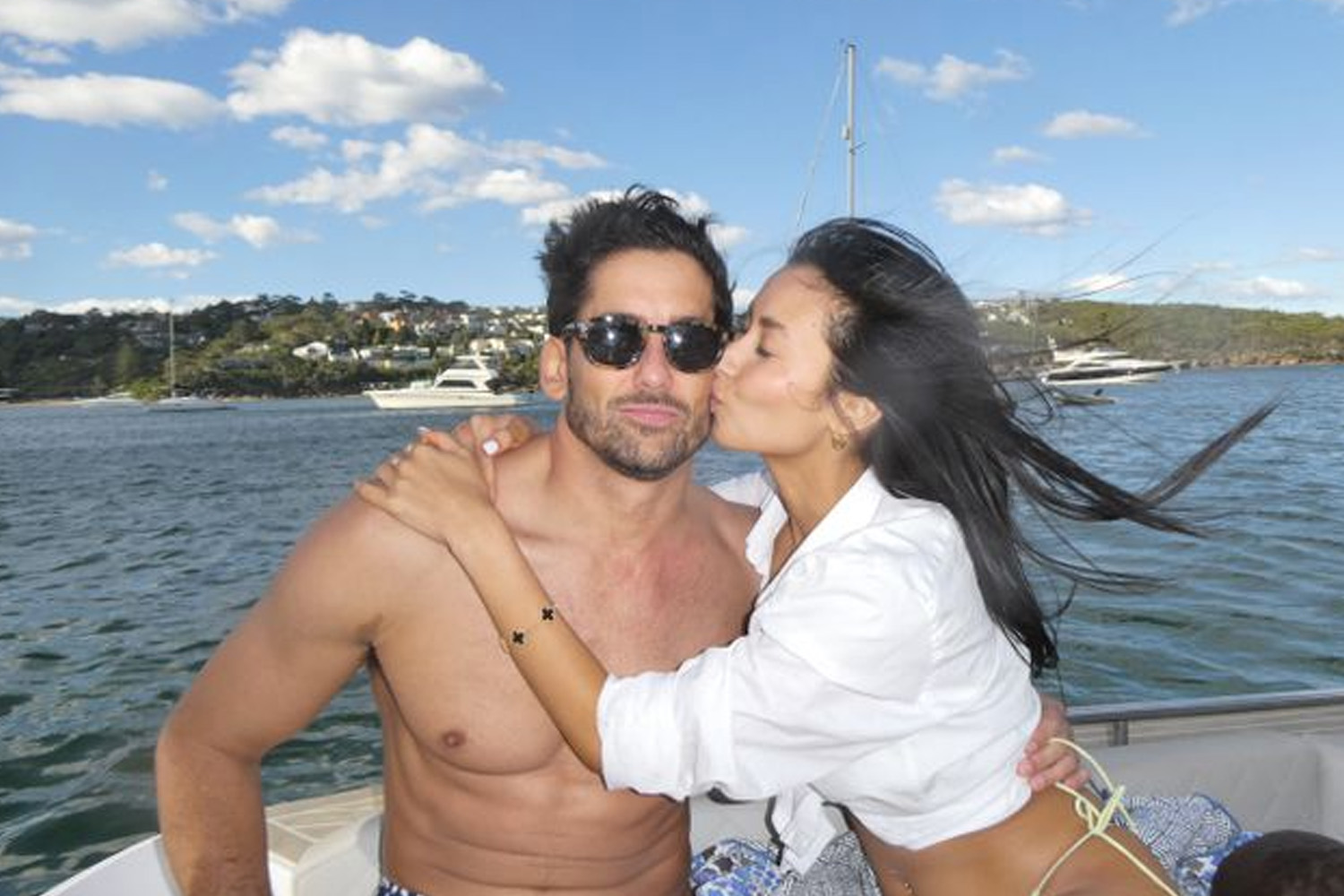 Man wearing sunglasses on a boat, with a woman kissing his cheek; both appear happy, with water and yachts in the background.