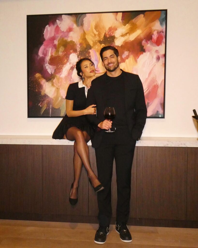 A couple dressed in black, with one seated on a counter, in front of a colorful abstract painting.
