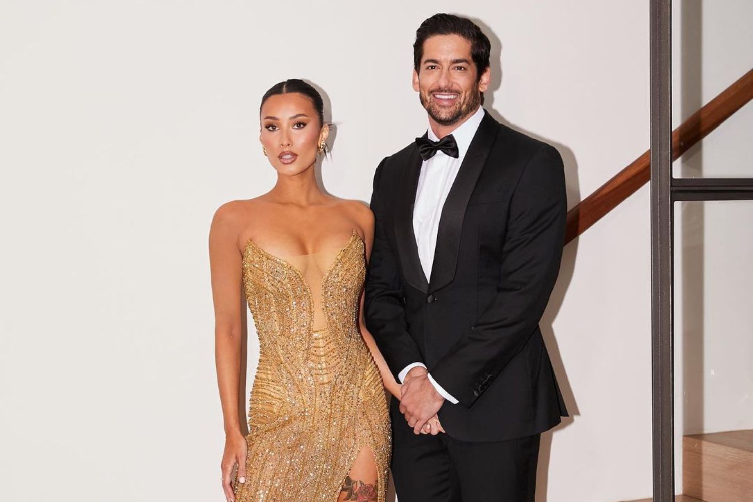 A couple posing elegantly; woman in gold dress, man in black tuxedo, both smiling.