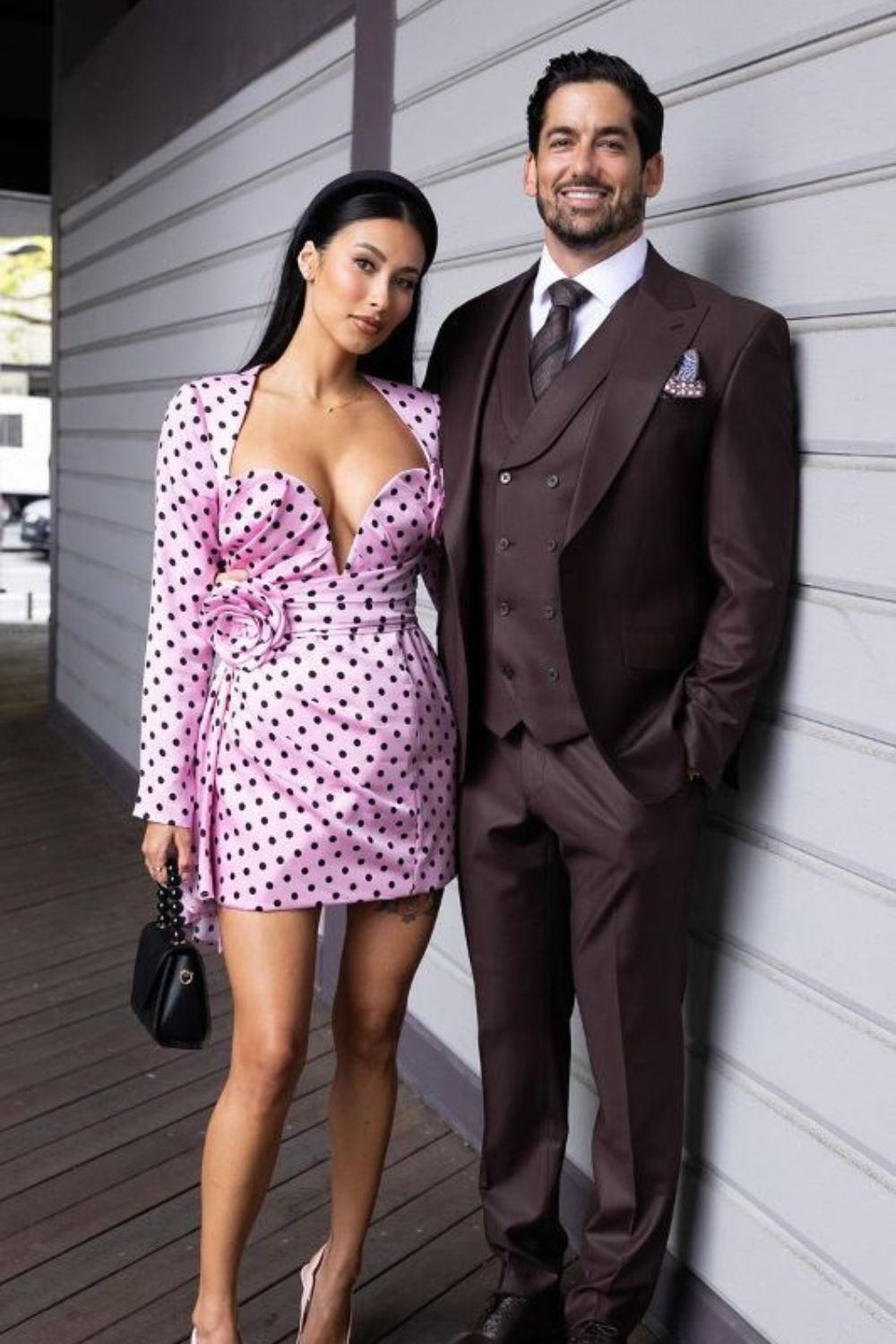 A woman in a pink polka dot dress stands next to a man in a brown suit, both smiling outside a building.