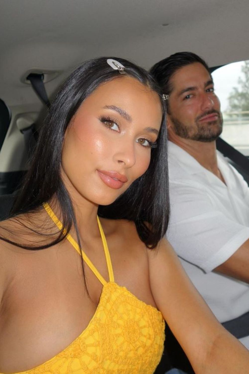 Young couple sitting in a car, woman in a yellow top takes selfie, man in a white shirt smiles in the background.