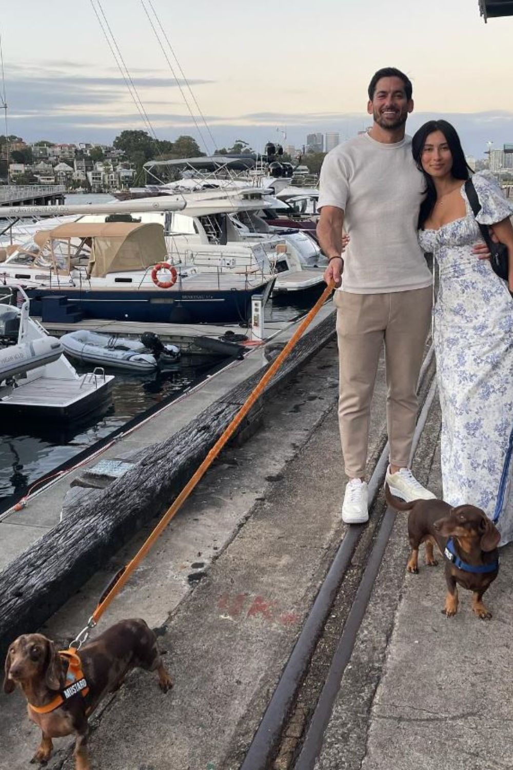 Couple walking two dachshunds along a marina with boats in the background.
