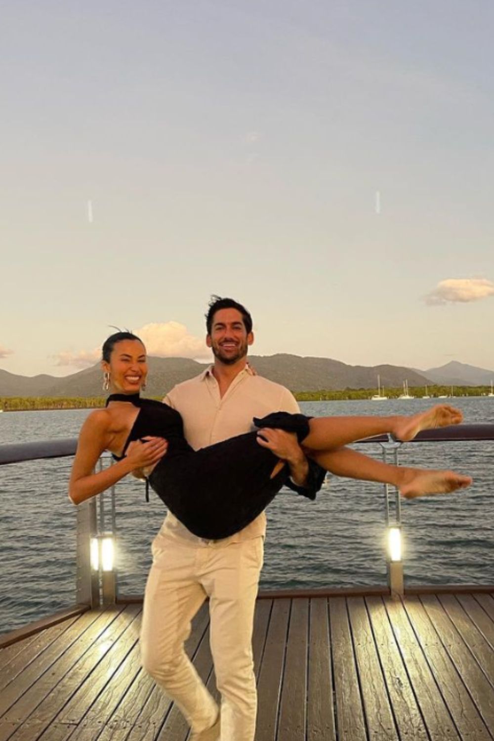 Man in white shirt holding woman in black dress; both smiling on a dock with water and mountains in the background.