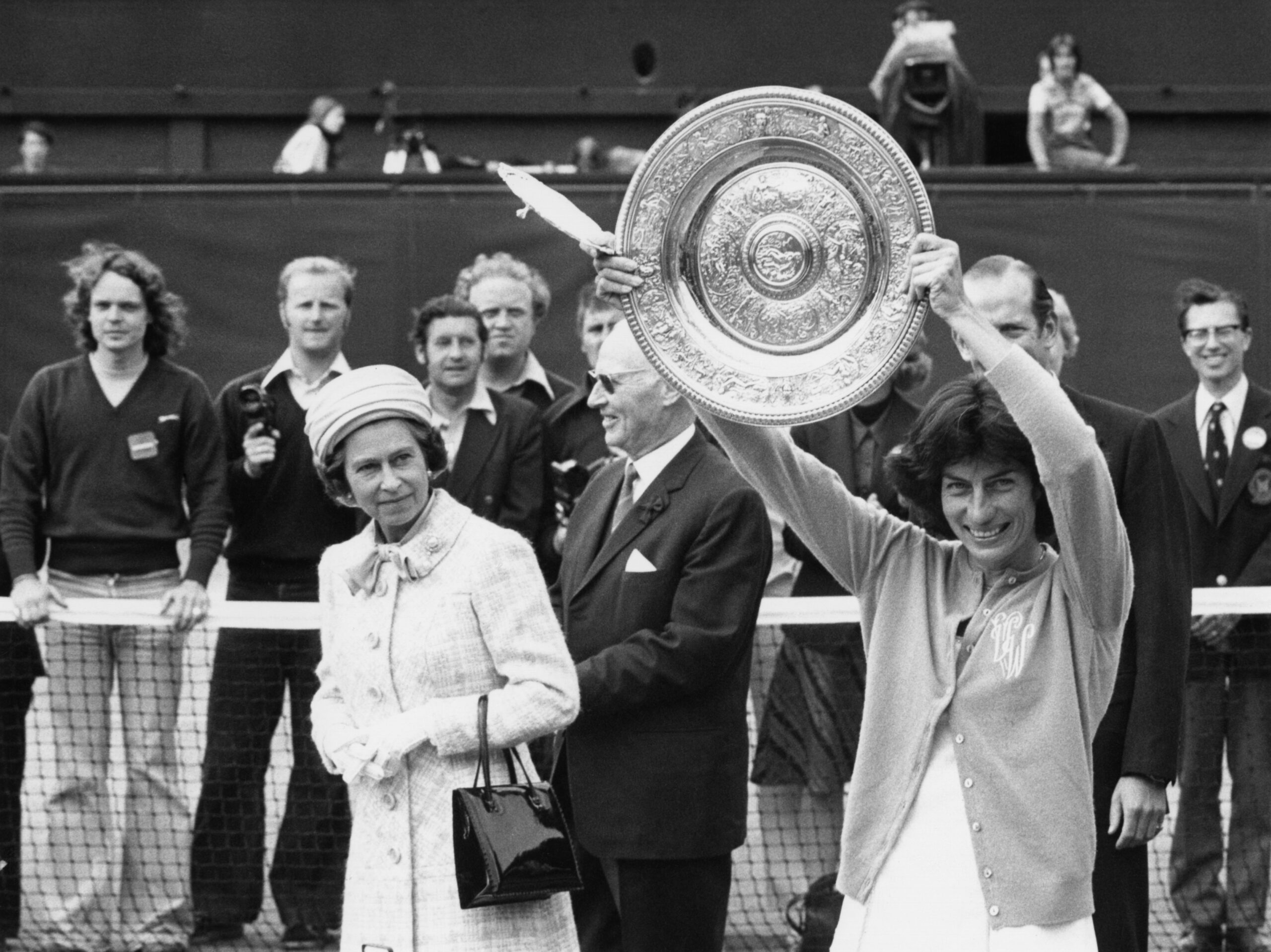 A woman holds a trophy aloft at a tennis event, with an elegantly dressed lady beside her and a crowd in the background.