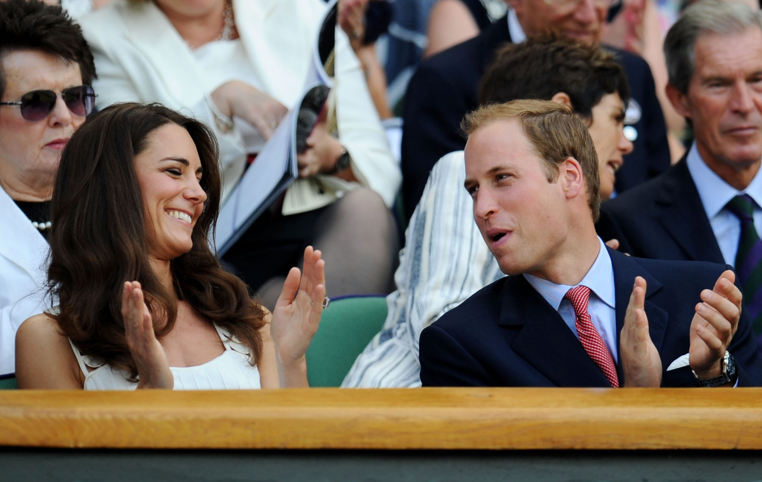 A woman in a white dress and a man in a suit applaud and smile while sitting in the audience at an event.