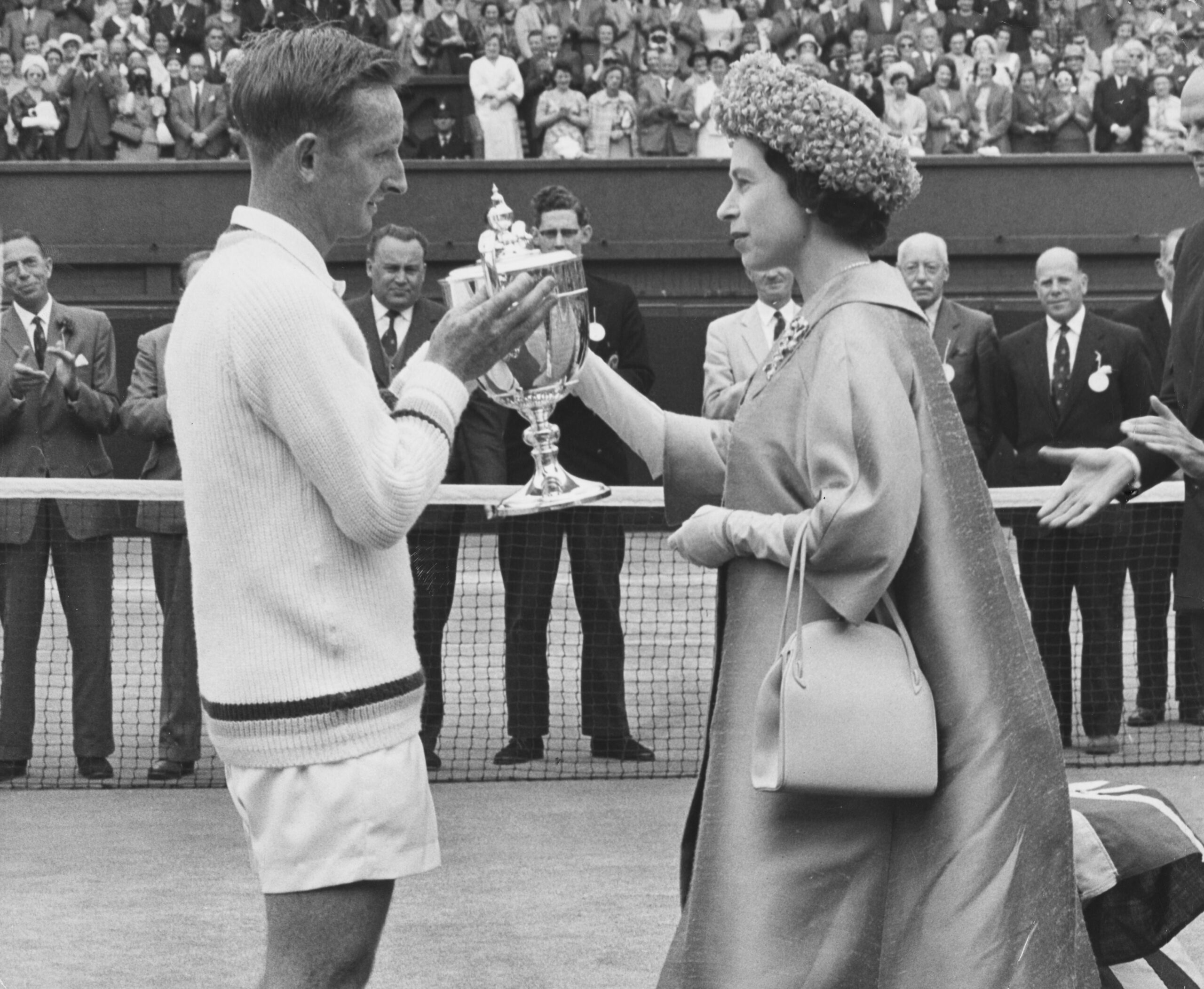 A man in tennis attire receives a trophy from a woman wearing a cape and hat on a tennis court, with a crowd watching.