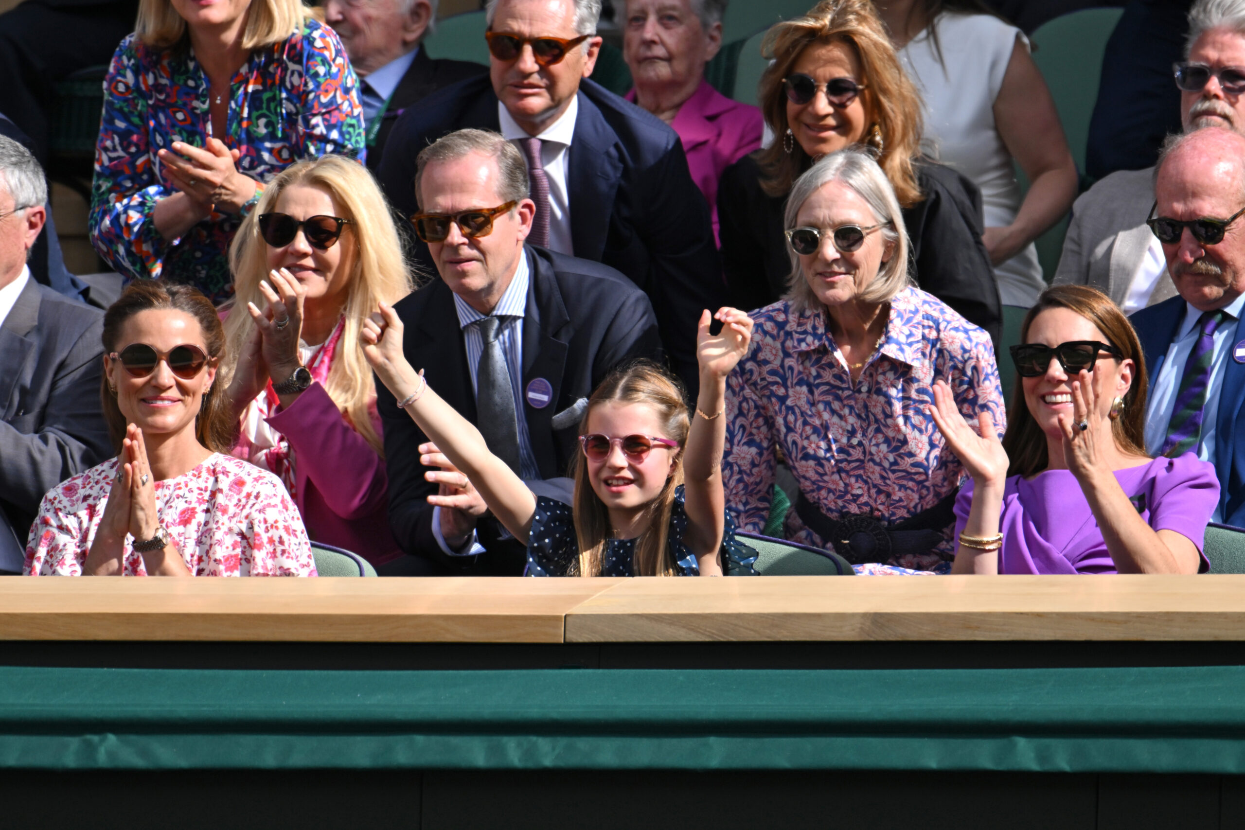 People in sunglasses, including a girl raising her arms, are clapping at a tennis event.