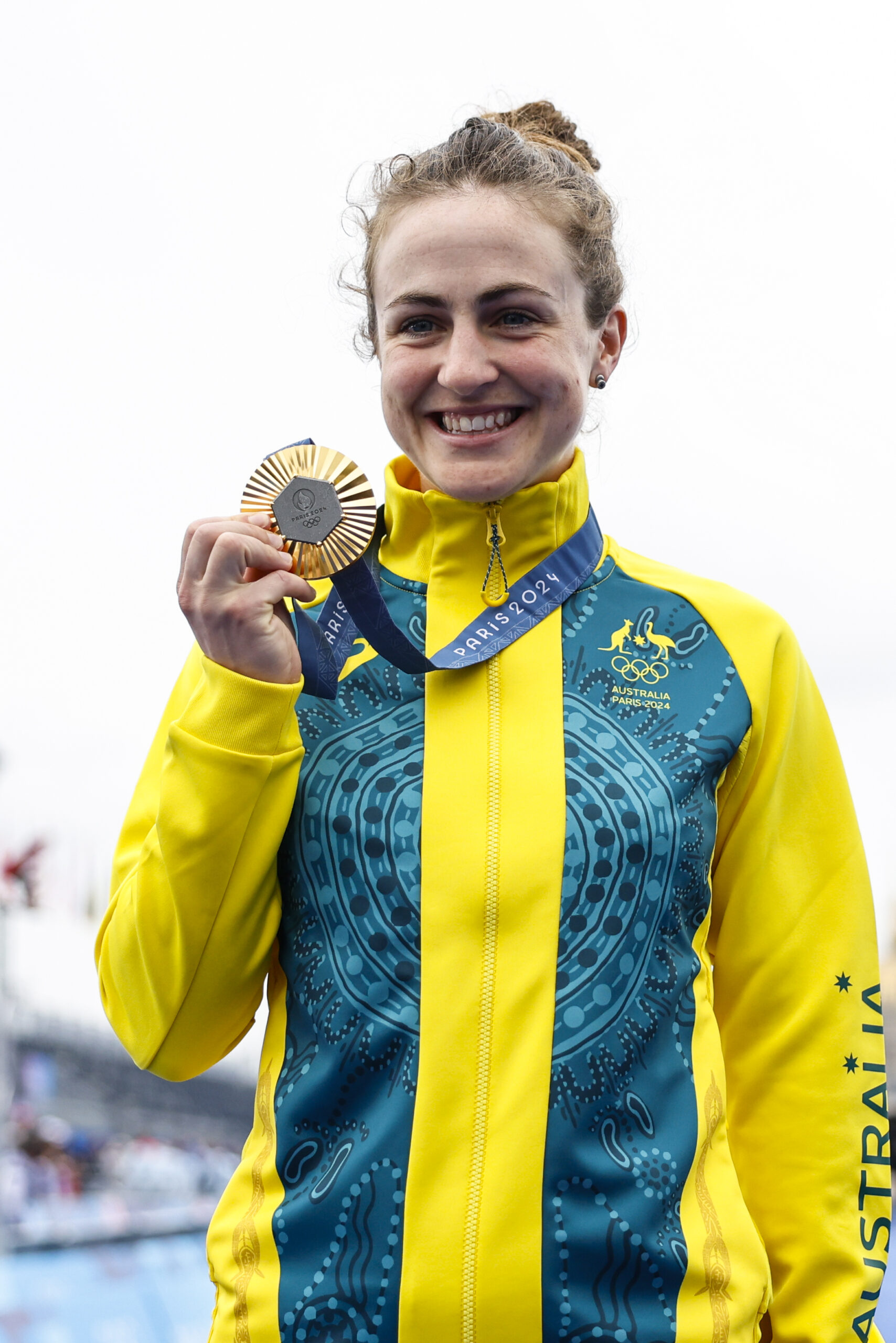 Australian athlete smiling, holding a silver medal, wearing a yellow and blue jacket with "PARIS 2024" lanyard.